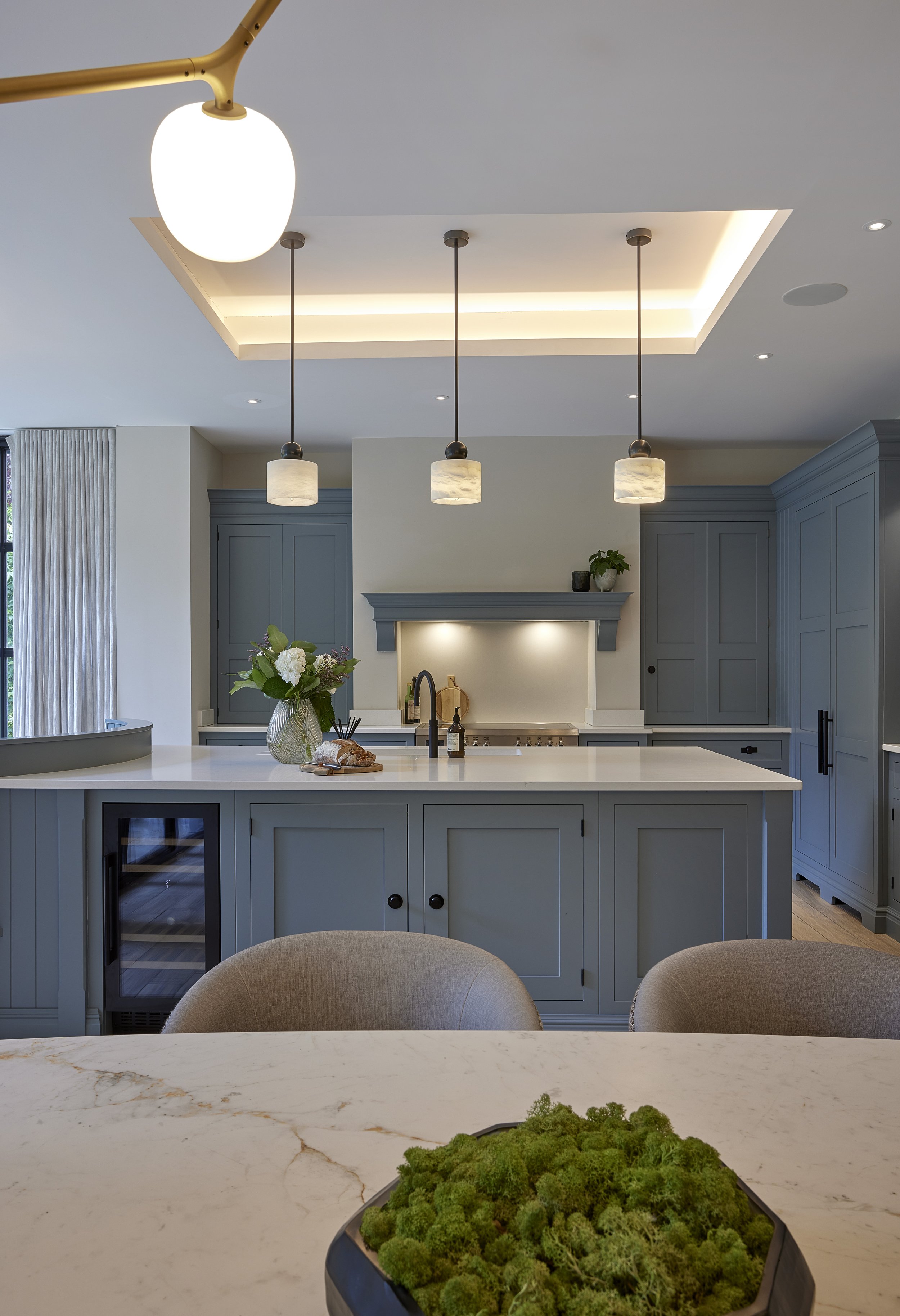 Modern kitchen with blue cabinetry, white countertops, pendant lighting, and a marble island with a green moss centerpiece.