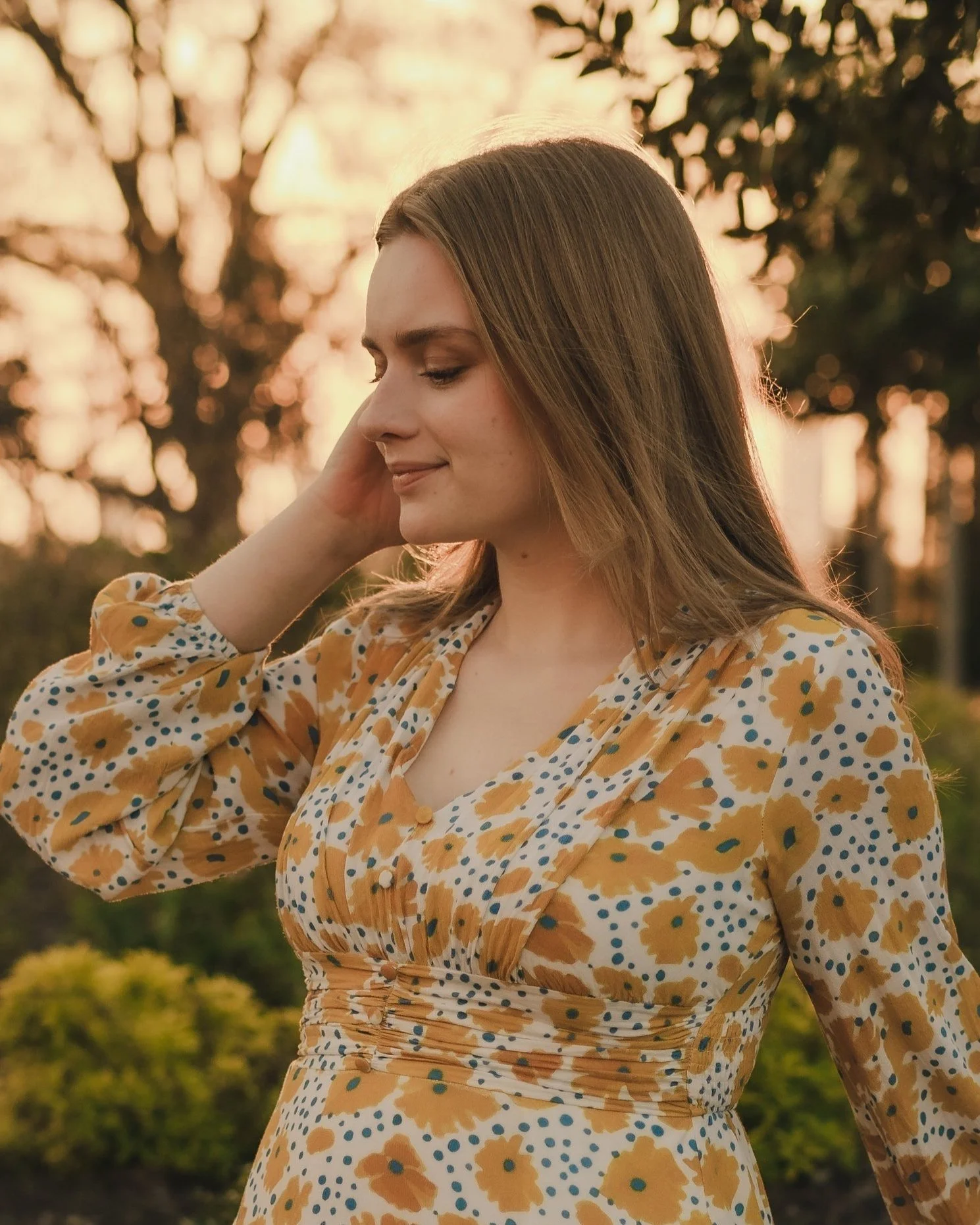 A woman with long blond hair smiling softly while standing outdoors at sunset, wearing a floral yellow, white, and blue dress.