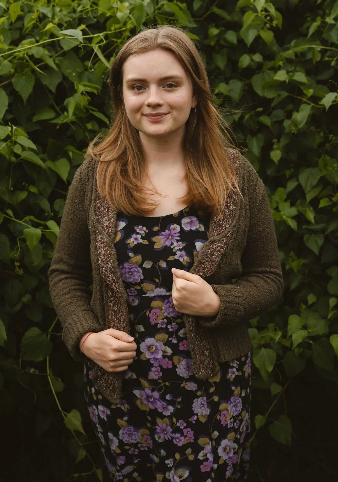 A young woman with long brown hair and fair skin standing outdoors in front of green leafy bushes. She is wearing a dark floral dress with purple and white flowers and a brown knitted cardigan. She is smiling softly at the camera.