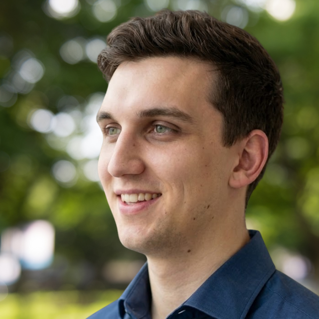 Man with short brown hair. He is smiling and wearing a dark blue shirt with a collar.