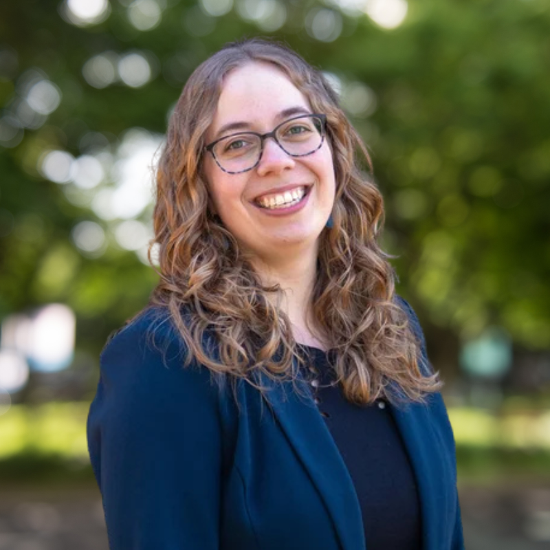 Women with blue blazer, medium length curly light brown hair and glasses.