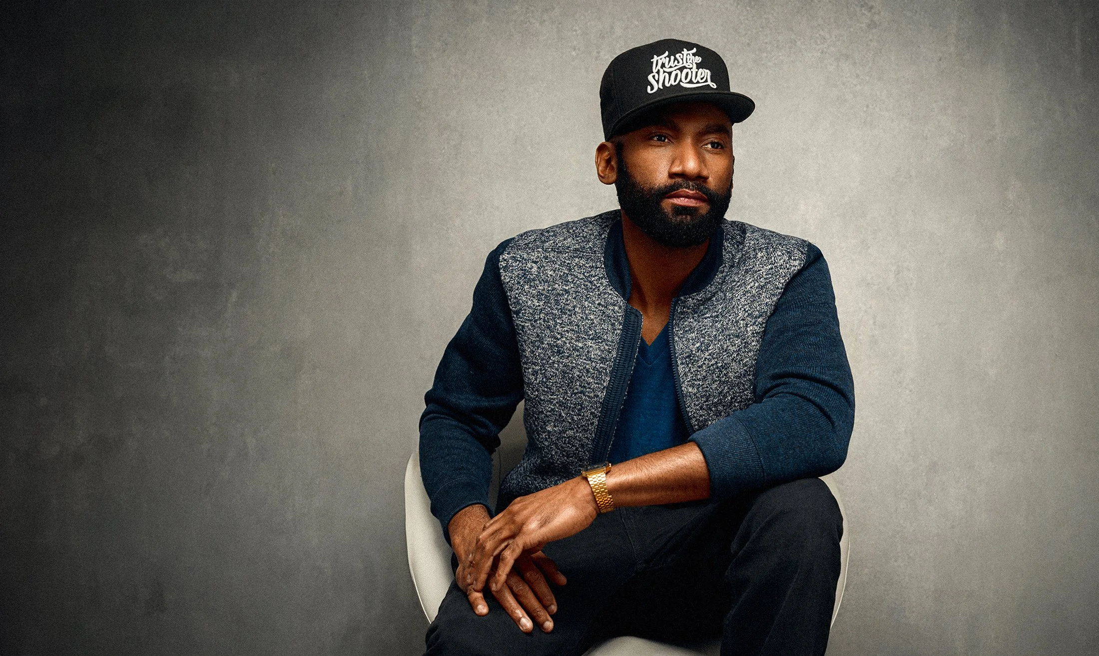 Un homme aux cheveux noirs et au regard sérieux, portant une casquette noire avec l'inscription "trust the shooter", une veste en jean et un bracelet doré, assis sur une chaise contre un mur gris.