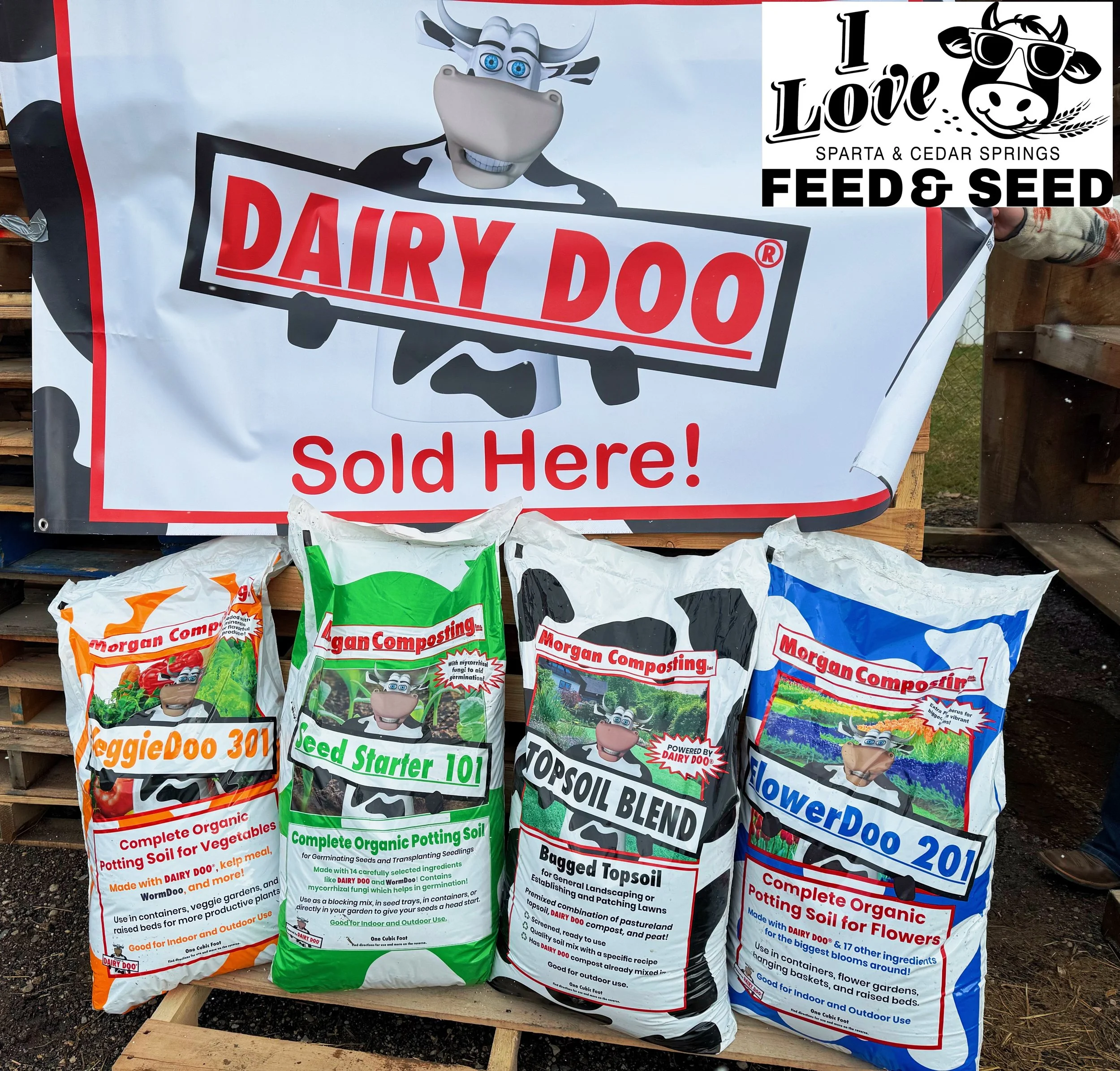 Display of garden soil products with a large sign that says 'Dairy Doo Sold Here!' and various bags labeled VeggieDoo 301, Seed Starter 101, Topsoil Blend, and FlowerDoo 201, all from Morgan Composting, at a farm or garden supply stand.