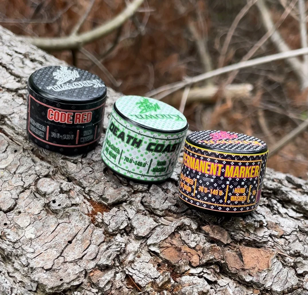 Three small, colorful containers labeled 'Code Red,' 'Death Coat,' and 'Permanent Marker' arranged on a tree log outdoors.