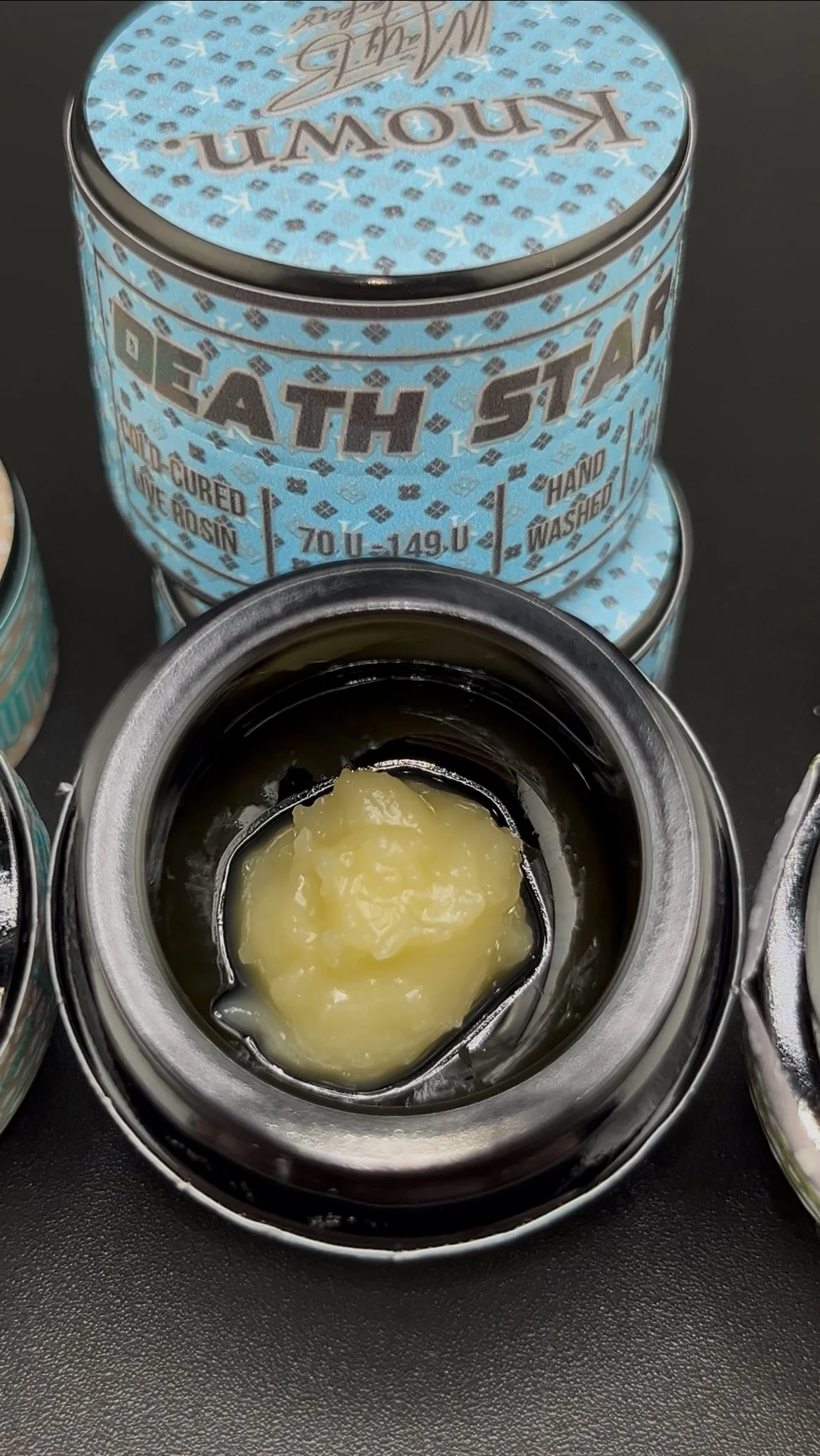 Open can of petroleum jelly with a thumb on the inner rim, surrounded by a blue tin labeled 'Death Star' with health and safety labels.