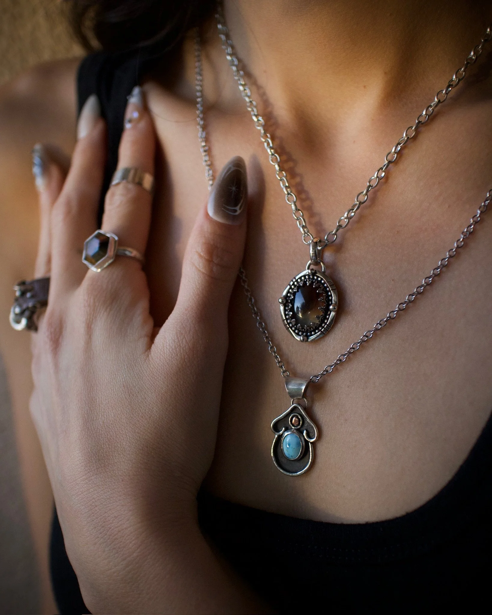 Two women wearing an assortment of silver jewelry, including necklaces, rings, and bracelets, with one woman wearing a white tank top and the other wearing a black zip-up hoodie.