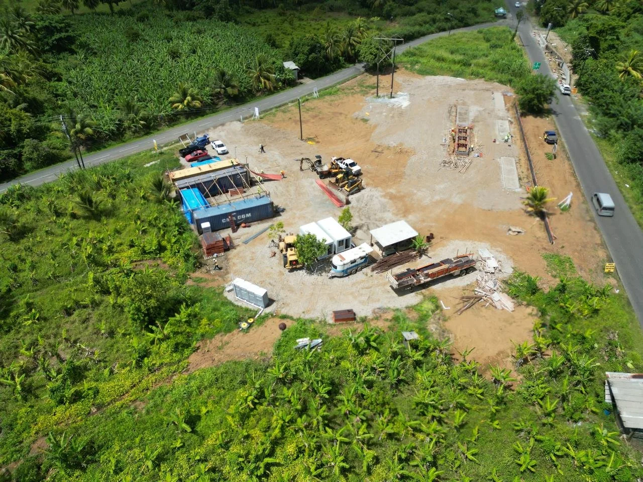 A construction site with machinery, vehicles, and materials surrounded by lush green trees and fields, with a road running along the edge.