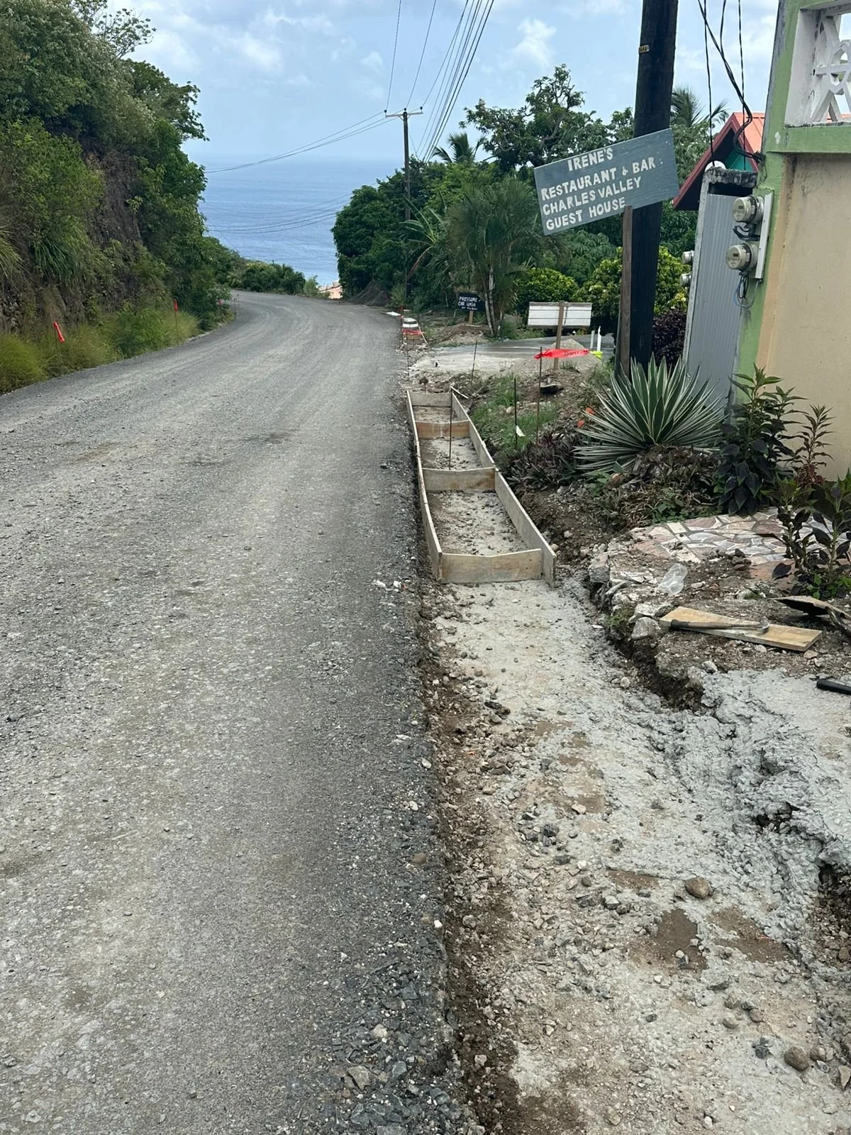Ongoing construction on a roadside with a gravel road, partial sidewalk, and signs for a restaurant named Irene's in a lush, tropical area with the ocean visible in the distance.