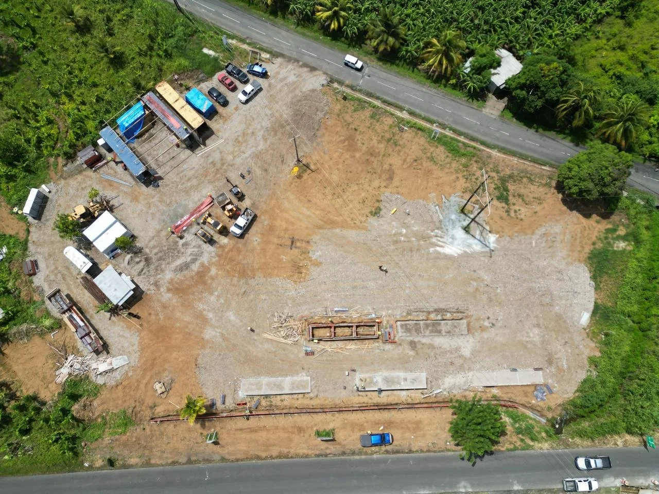 Aerial view of a construction site with building materials, vehicles, and equipment, surrounded by green trees and near a paved road.