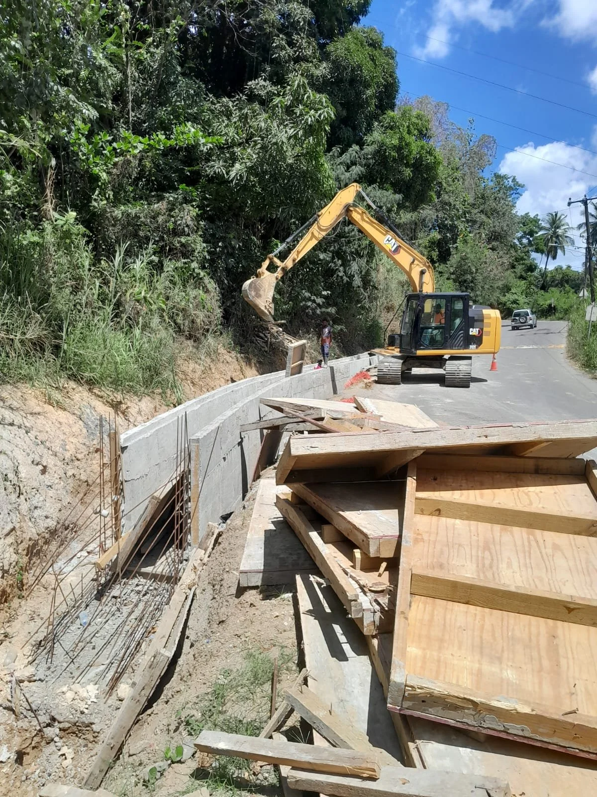 Construction site on a hillside with a yellow excavator working on a retaining wall. There are wooden and metal supports, a partially built concrete wall, and a person standing near the construction area. The scene is set on a rural paved road with t