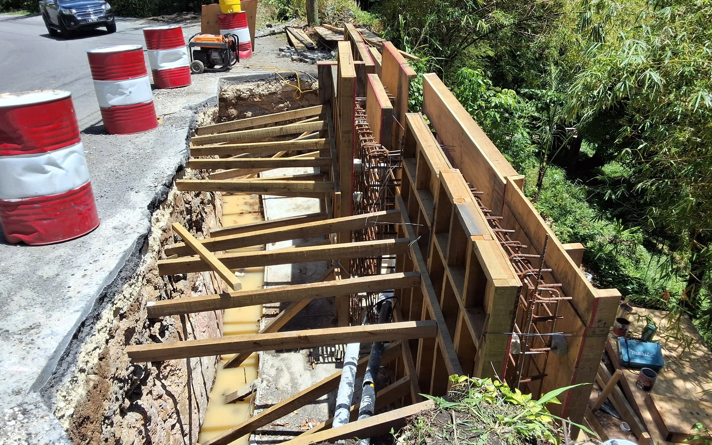 Construction site of a retaining wall with wooden and metal supports, adjacent to a road with traffic barrels and a parked vehicle, surrounded by greenery.