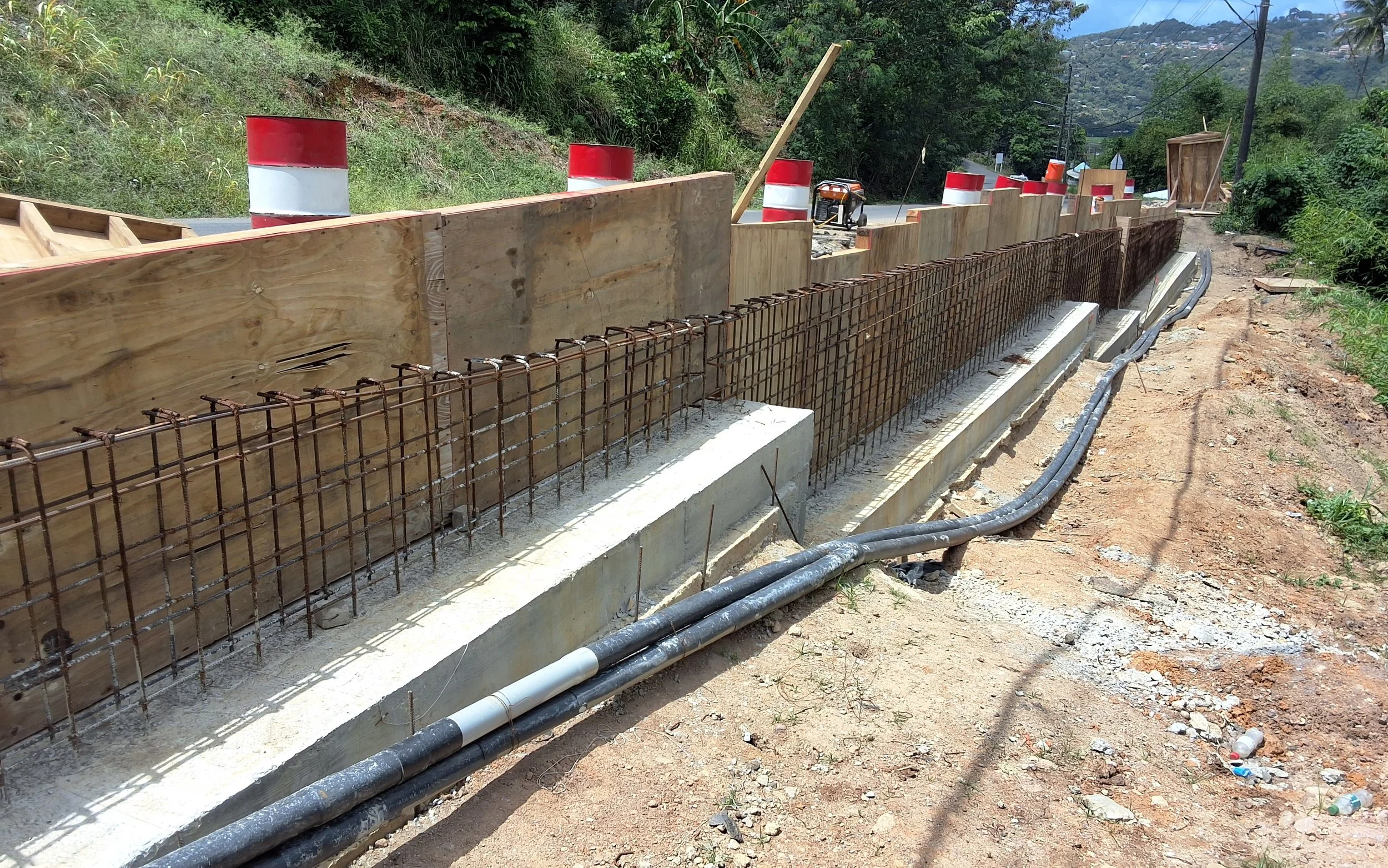 Construction site with a partially built concrete barrier reinforced with metal rebar, wooden formwork, and orange and white traffic barrels along a roadway with greenery and hills in the background.