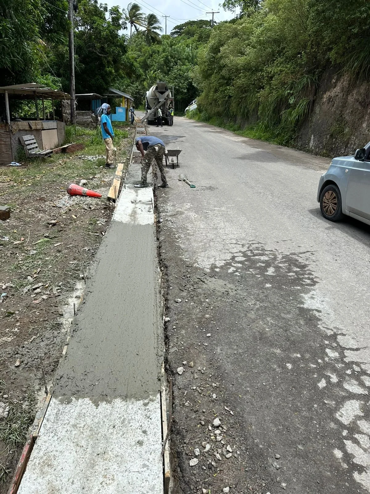 Workers are pouring concrete for a sidewalk along a rural road with trees and houses in the background.