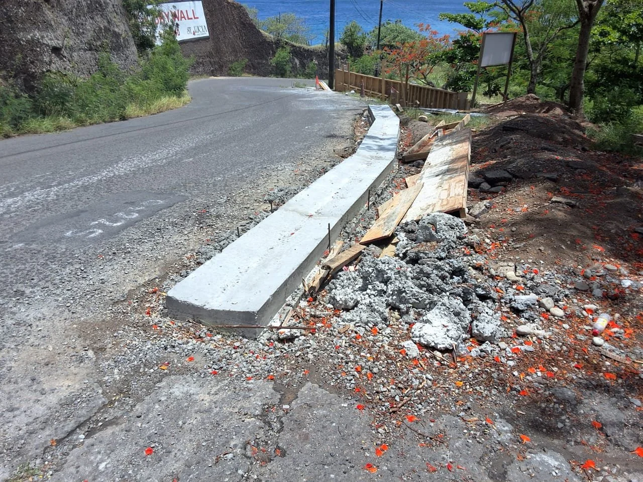 Construction site on a curving road with a new concrete curb being installed, piles of dirt and debris, a wooden fence on the right, trees, and signs in the background.