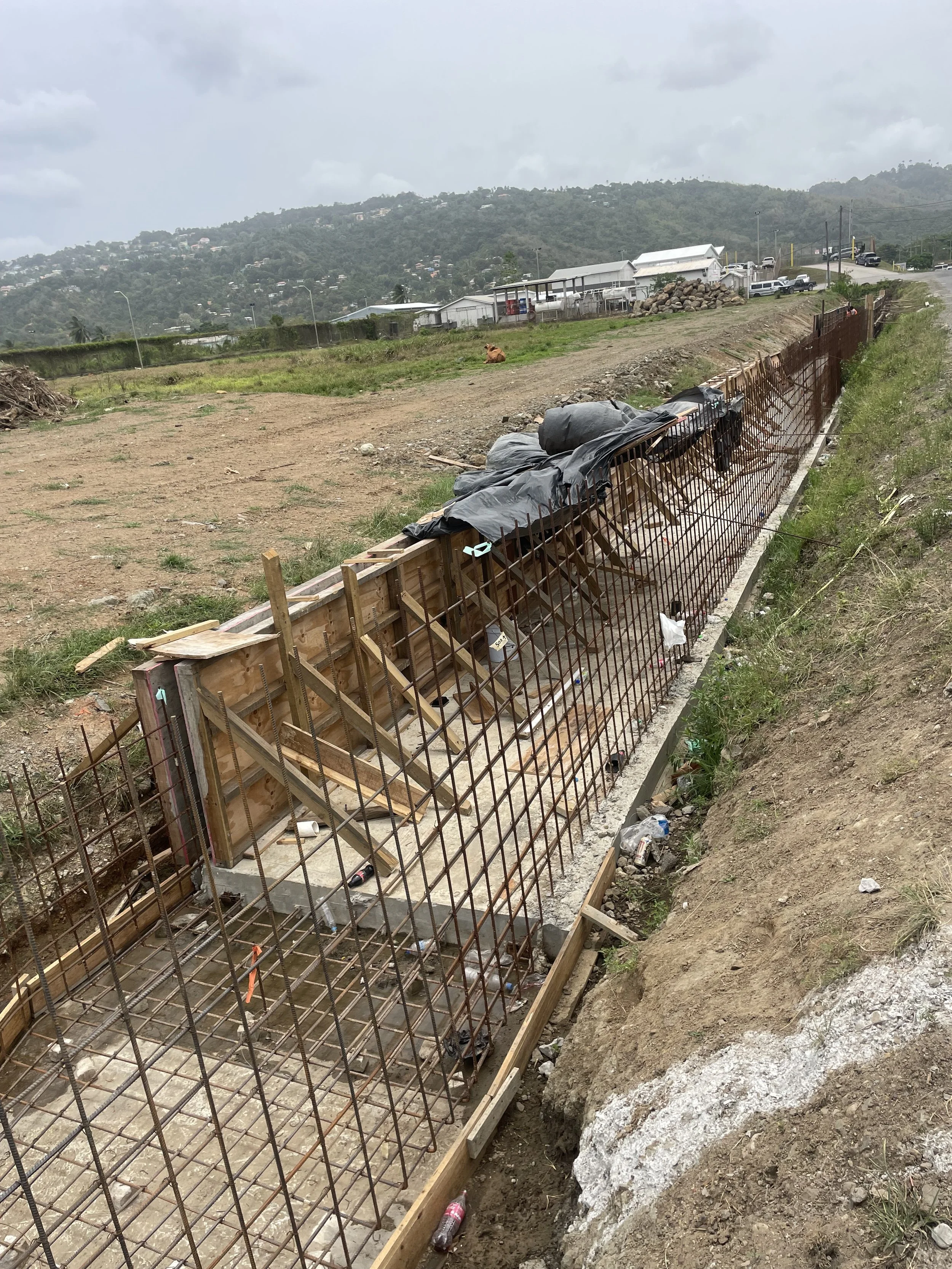 Construction site with a rebar fence and concrete foundation, hill and cloudy sky in the background.