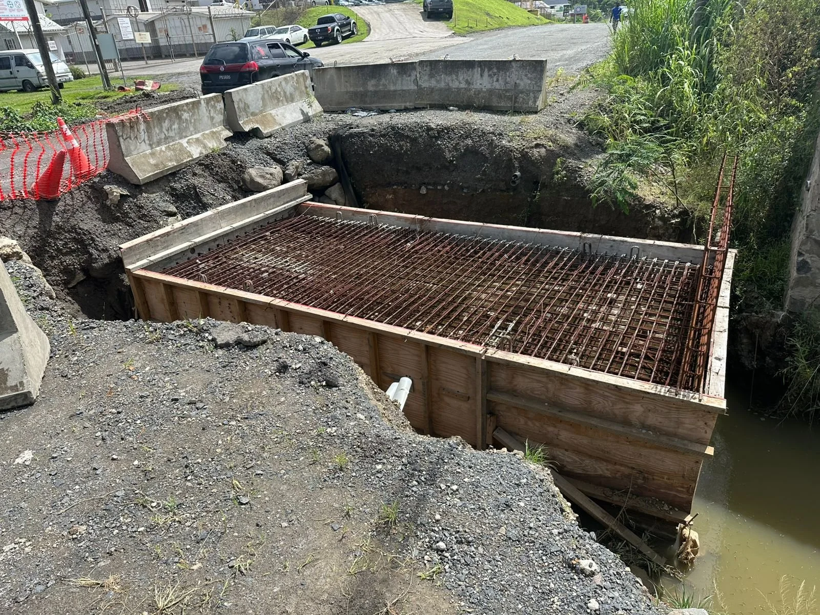 Construction site with a wooden form and steel rebar structure in a water-filled excavation, surrounded by dirt and gravel, with concrete barriers and parked cars in the background.