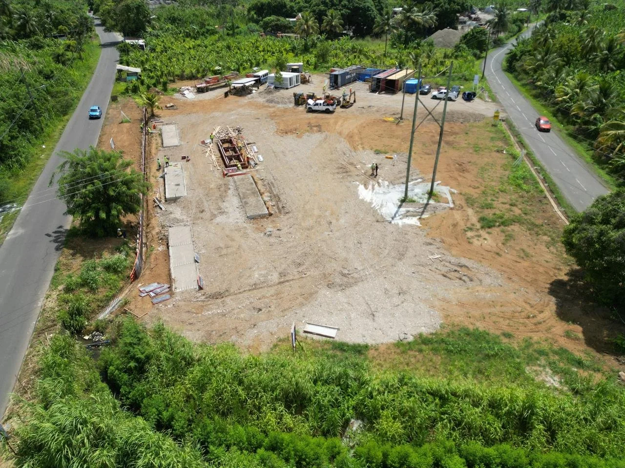 Aerial view of a construction site with machinery, containers, and workers, surrounded by greenery and with roads on either side.