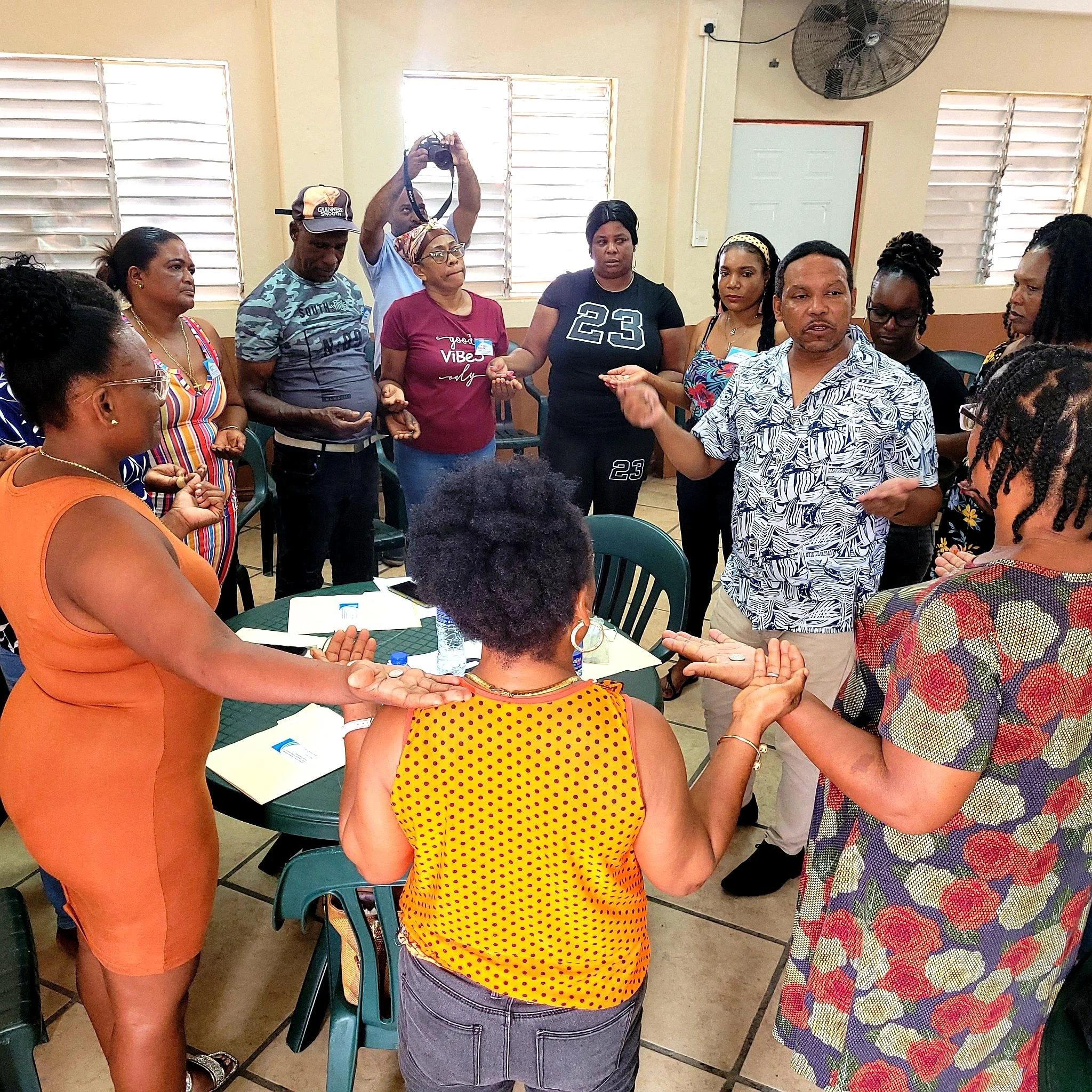 A diverse group of people hold hands in a circle during a meeting or workshop in a room with beige walls and windows, with some participants speaking and others listening attentively.