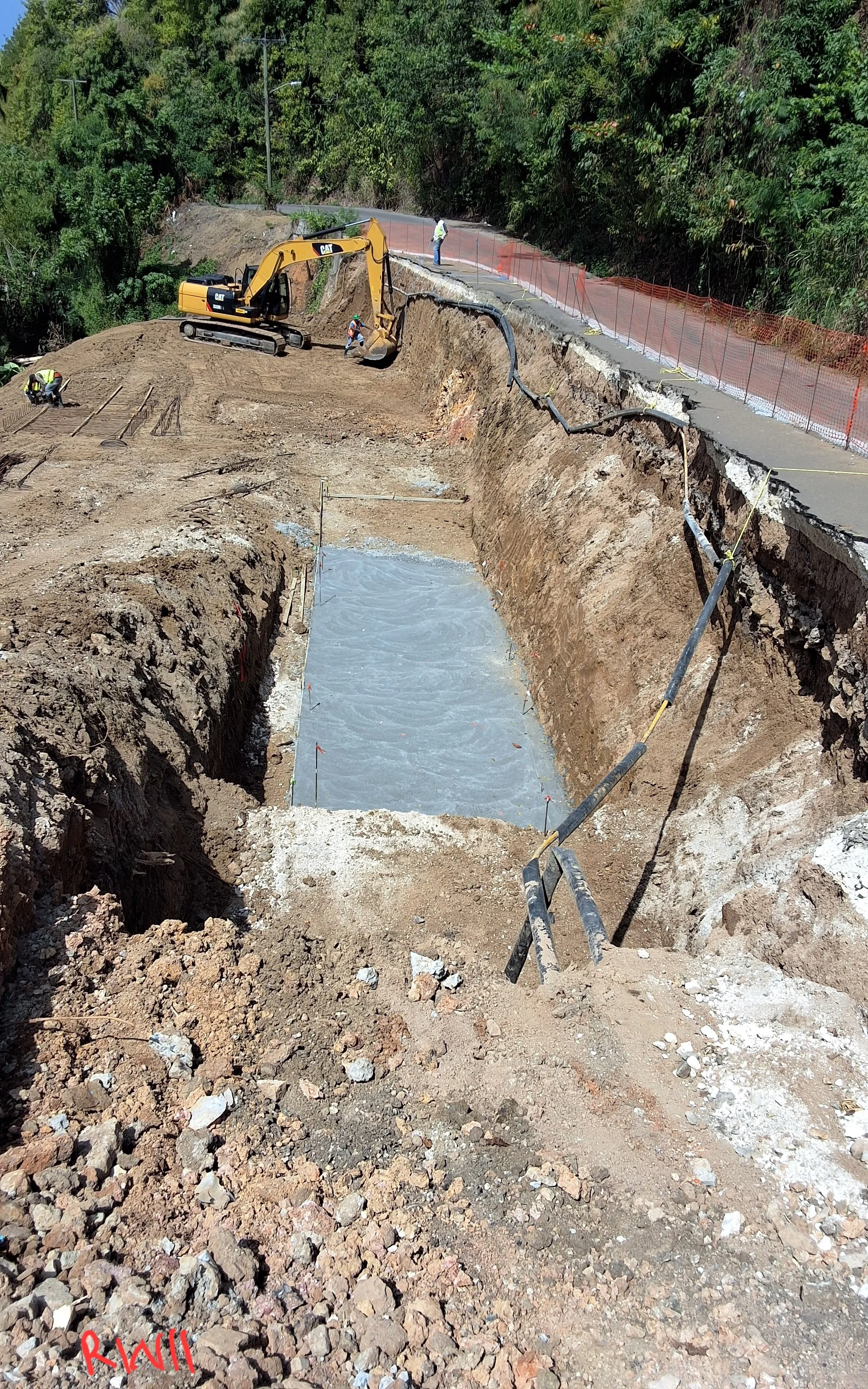 Construction site on a hillside where workers are building a retaining wall or foundation. Equipment includes an excavator and shovels, with a section of road above and a trench with concrete reinforcement below.