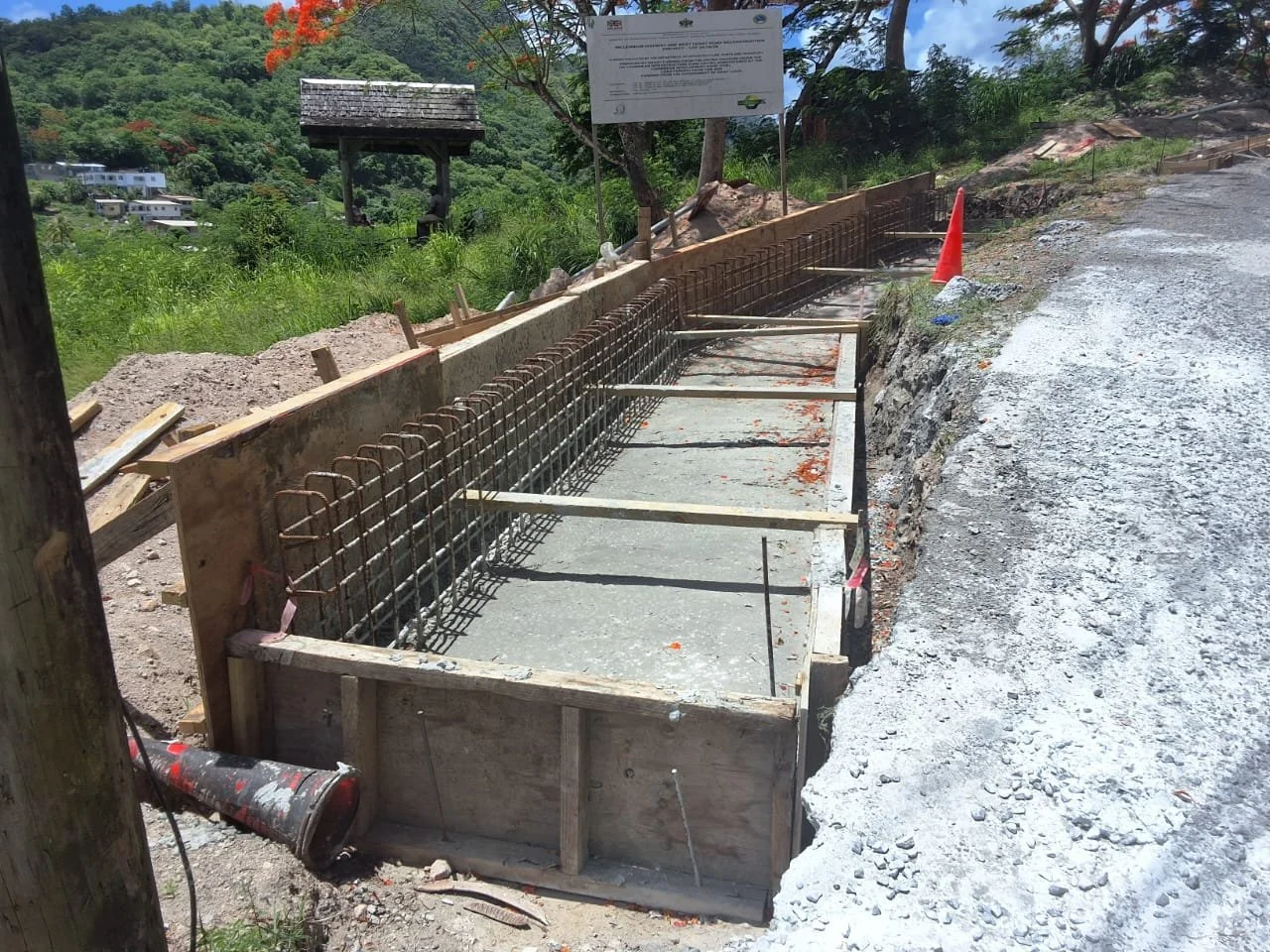 Construction site with rebar and concrete pavement in progress along a dirt road, with a traffic cone, trees, and a hillside in the background.