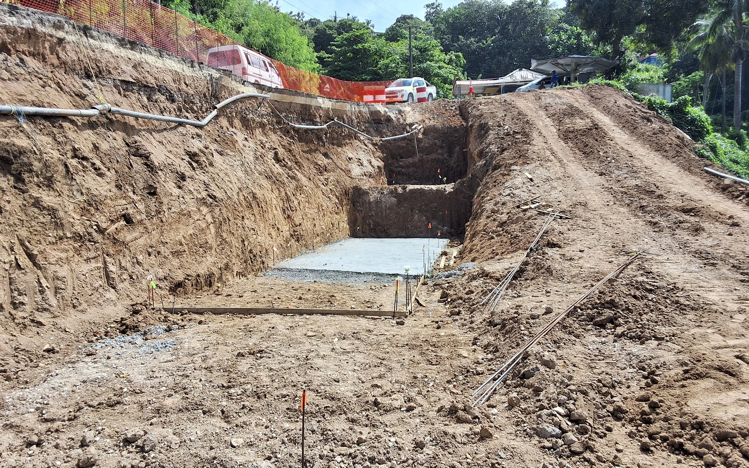 Construction site showing an excavated area with concrete foundation, metal rebar, and construction flags, with vehicles and trees in the background.