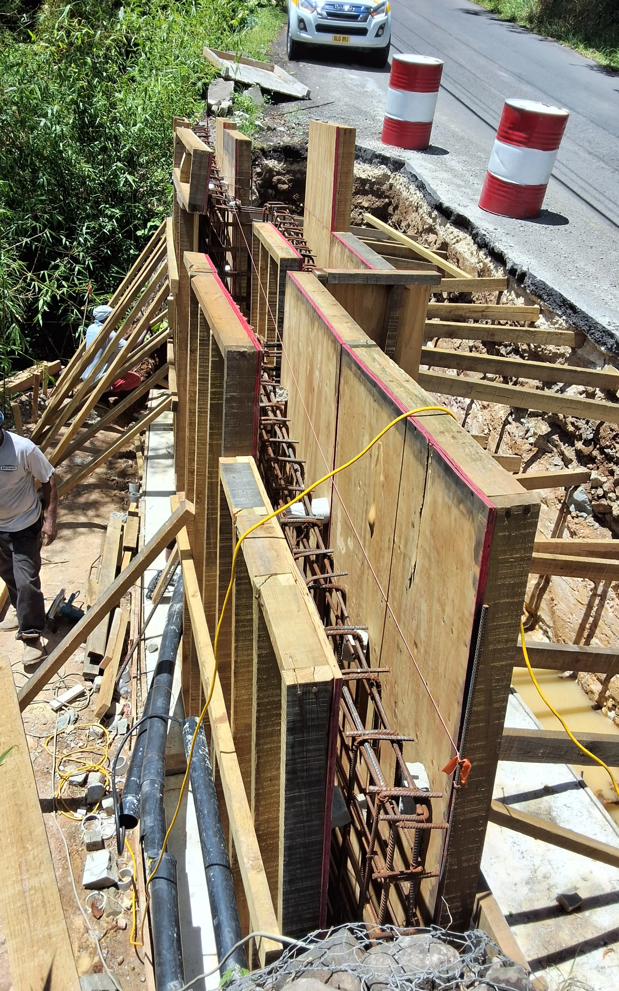 Construction workers building a concrete wall with wooden formwork and reinforcing steel bars, next to a rural road with traffic barrels and an approaching vehicle.