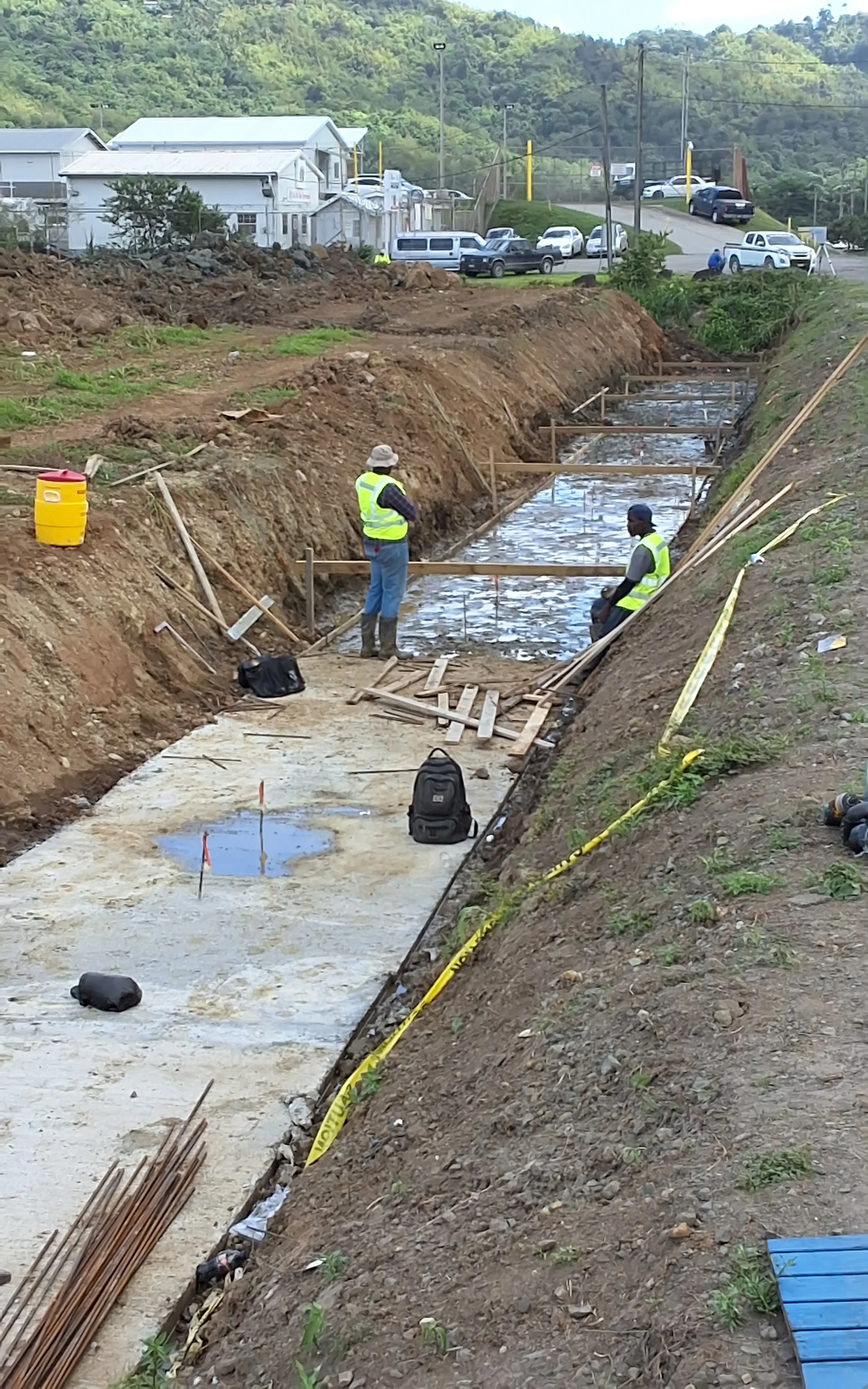 Construction workers building a concrete structure in a trench, surrounded by construction tools and materials, with a hilly area and parked cars in the background.