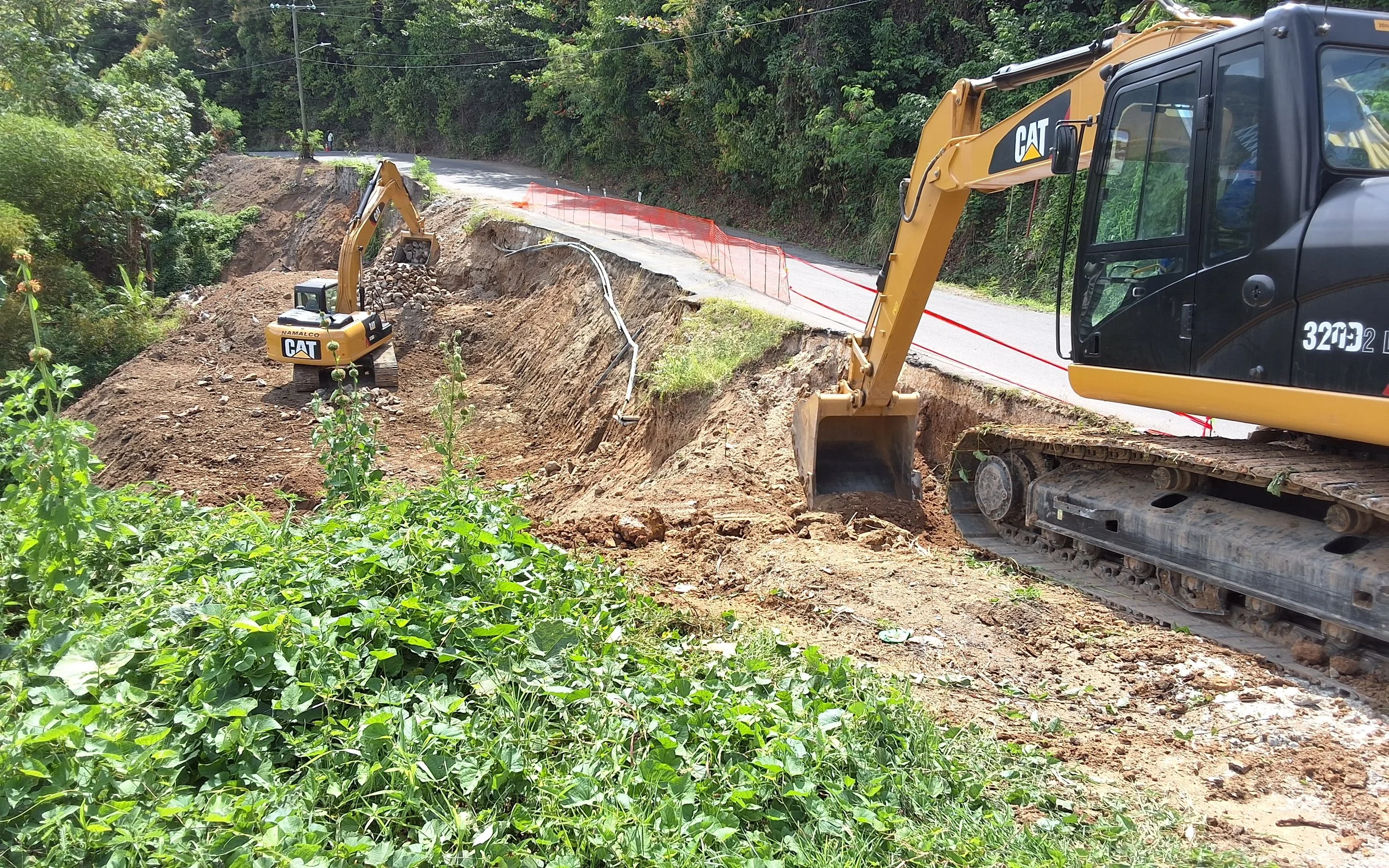 Two yellow Caterpillar excavators working on a slope beside a road, with green trees and orange safety netting in the background.