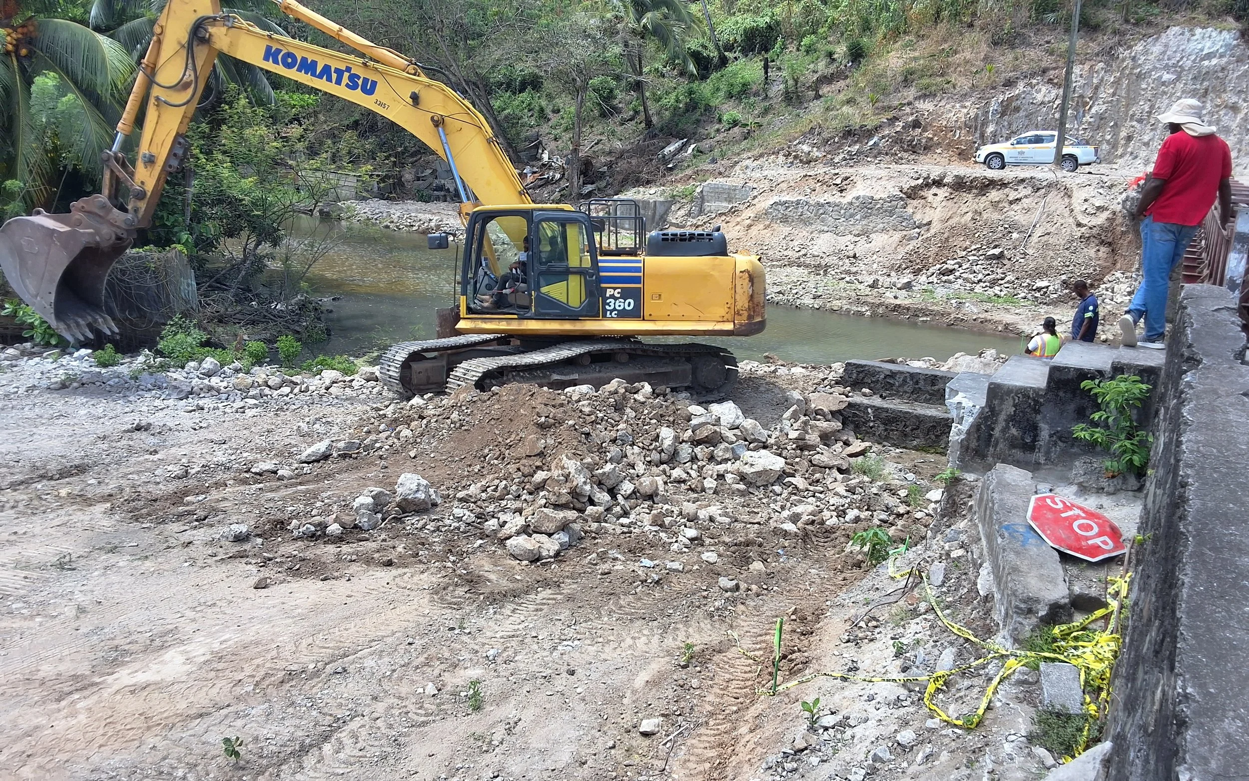 Construction site by a river with a Komatsu excavator moving dirt and rocks, several workers standing nearby, and a parked police vehicle on the hillside.