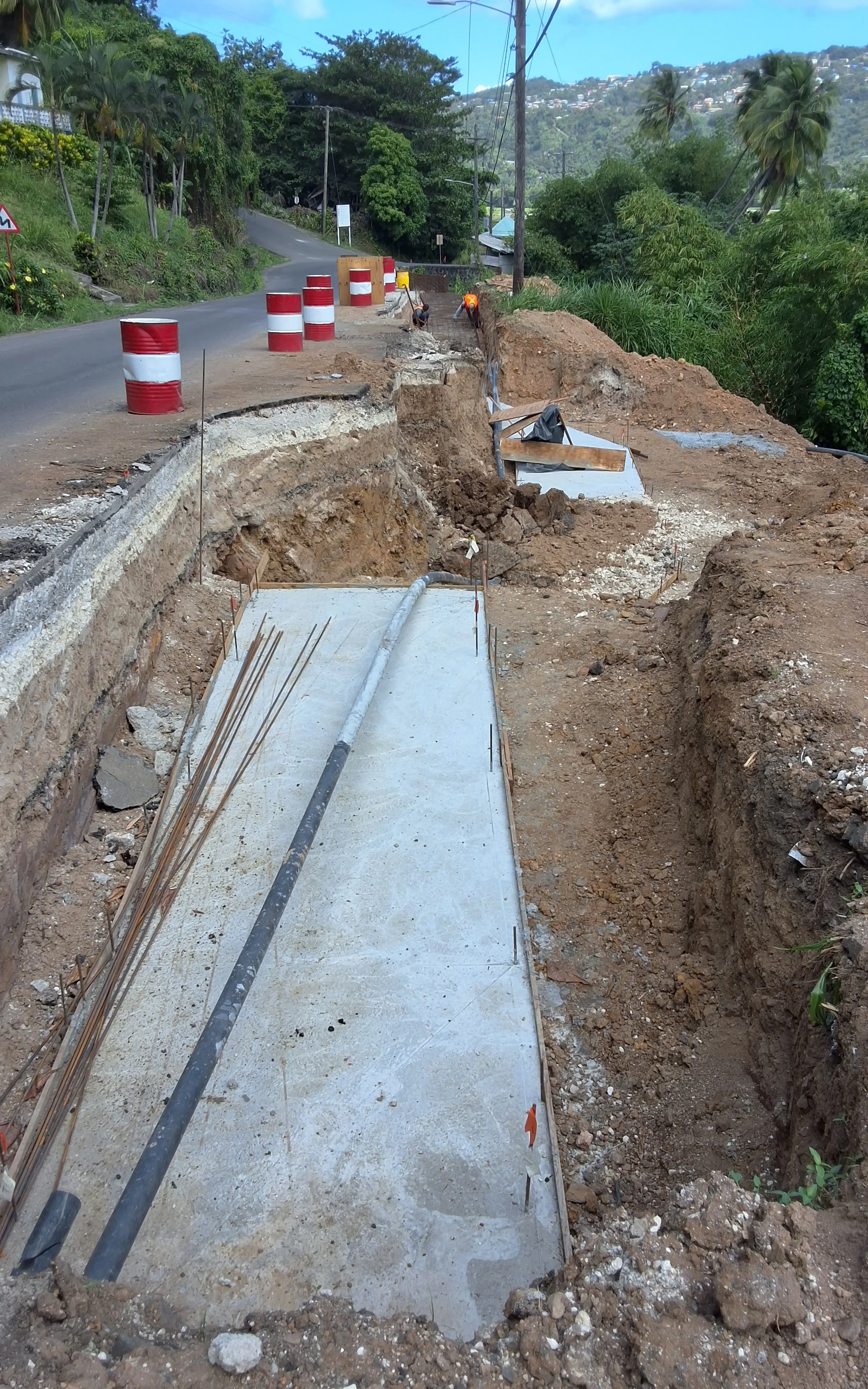 Construction workers working on a road with cement foundation, orange and white barrels separating the construction zone from the road, and a hilly green landscape in the background.