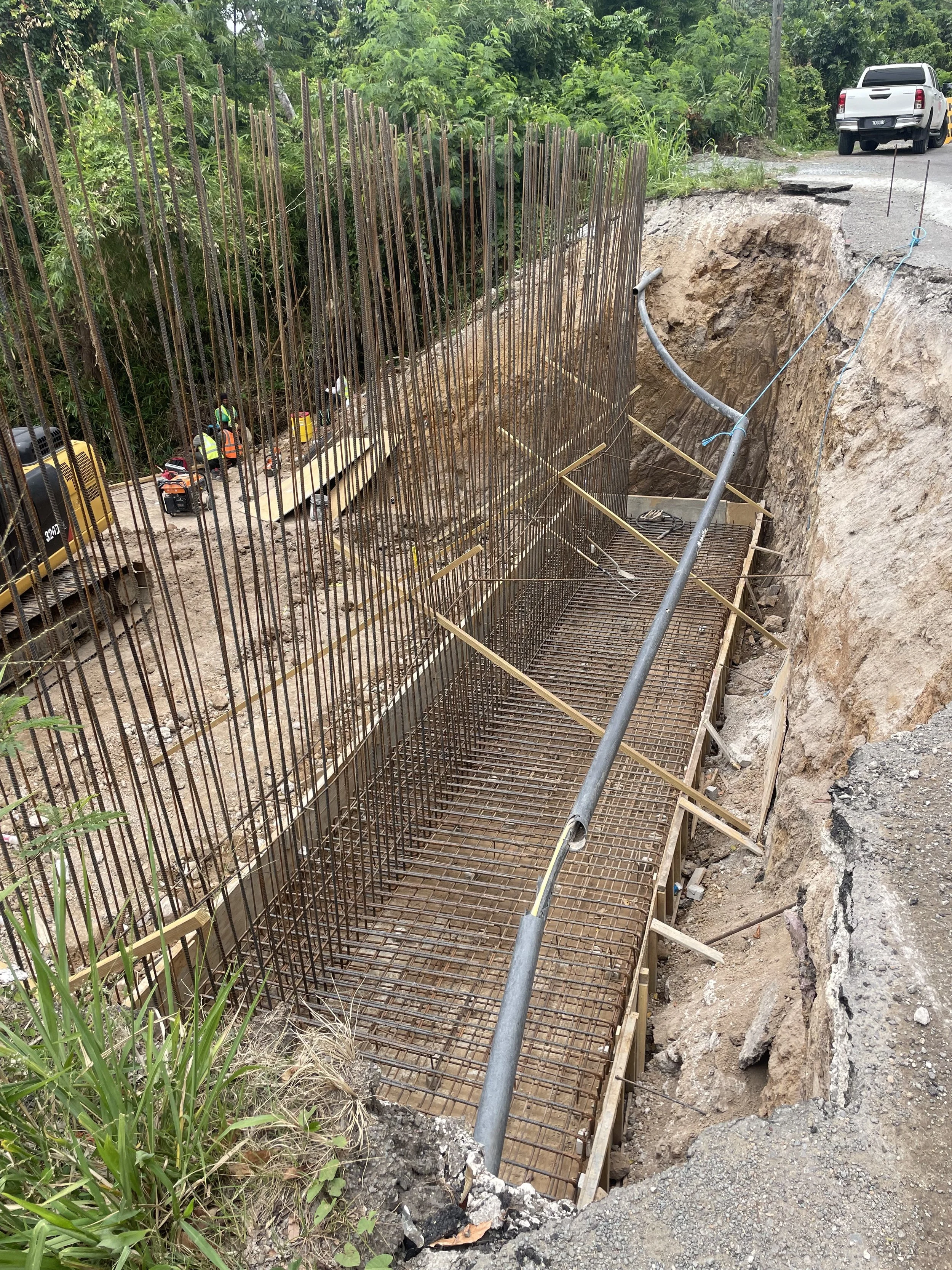 Construction site showing rebar framework and a pipe for a bridge or structure, with workers and machinery in the background, surrounded by greenery.