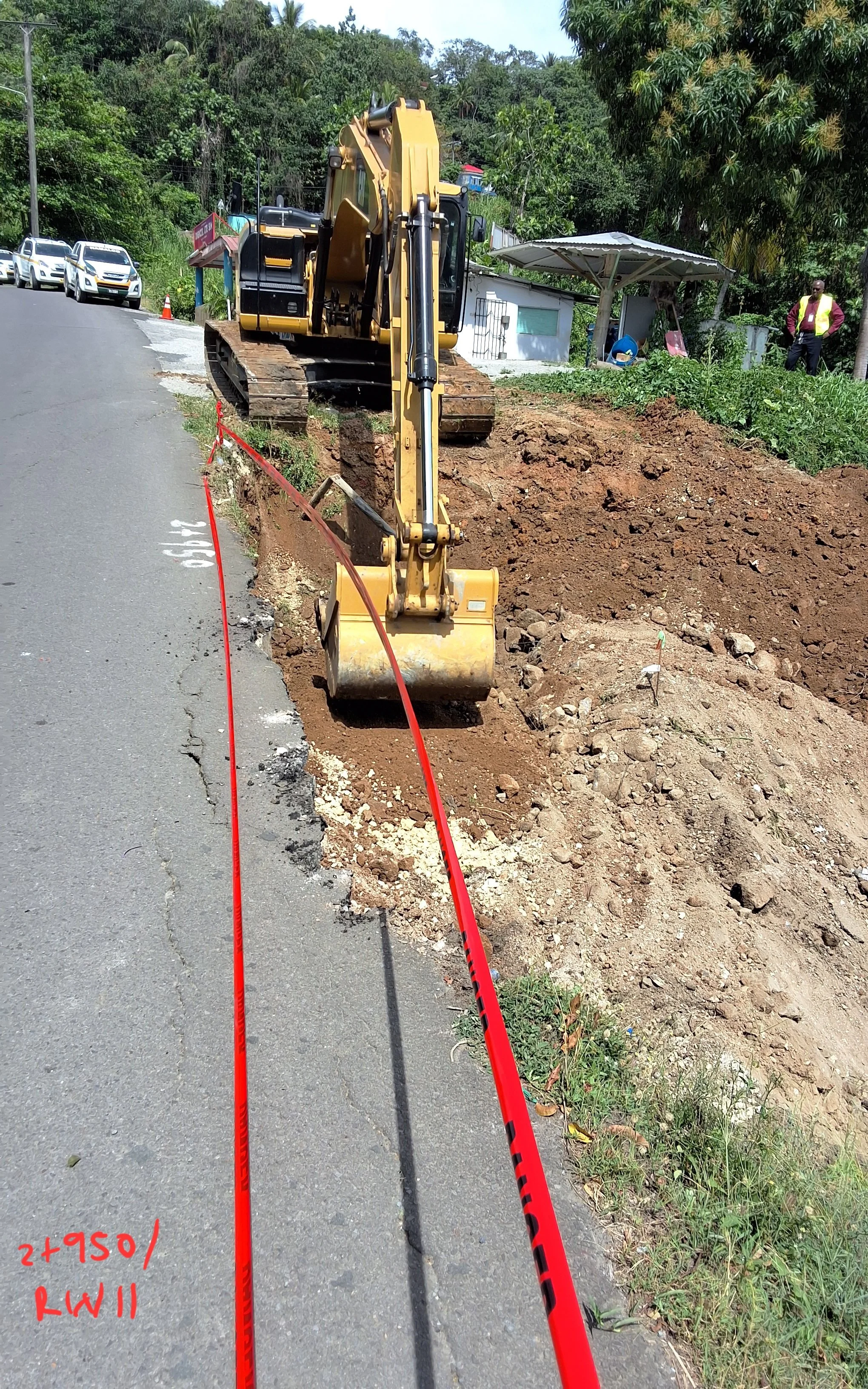 Construction site on a road with an excavator digging into the ground, orange safety tape marking the area, and a few construction workers in the background.