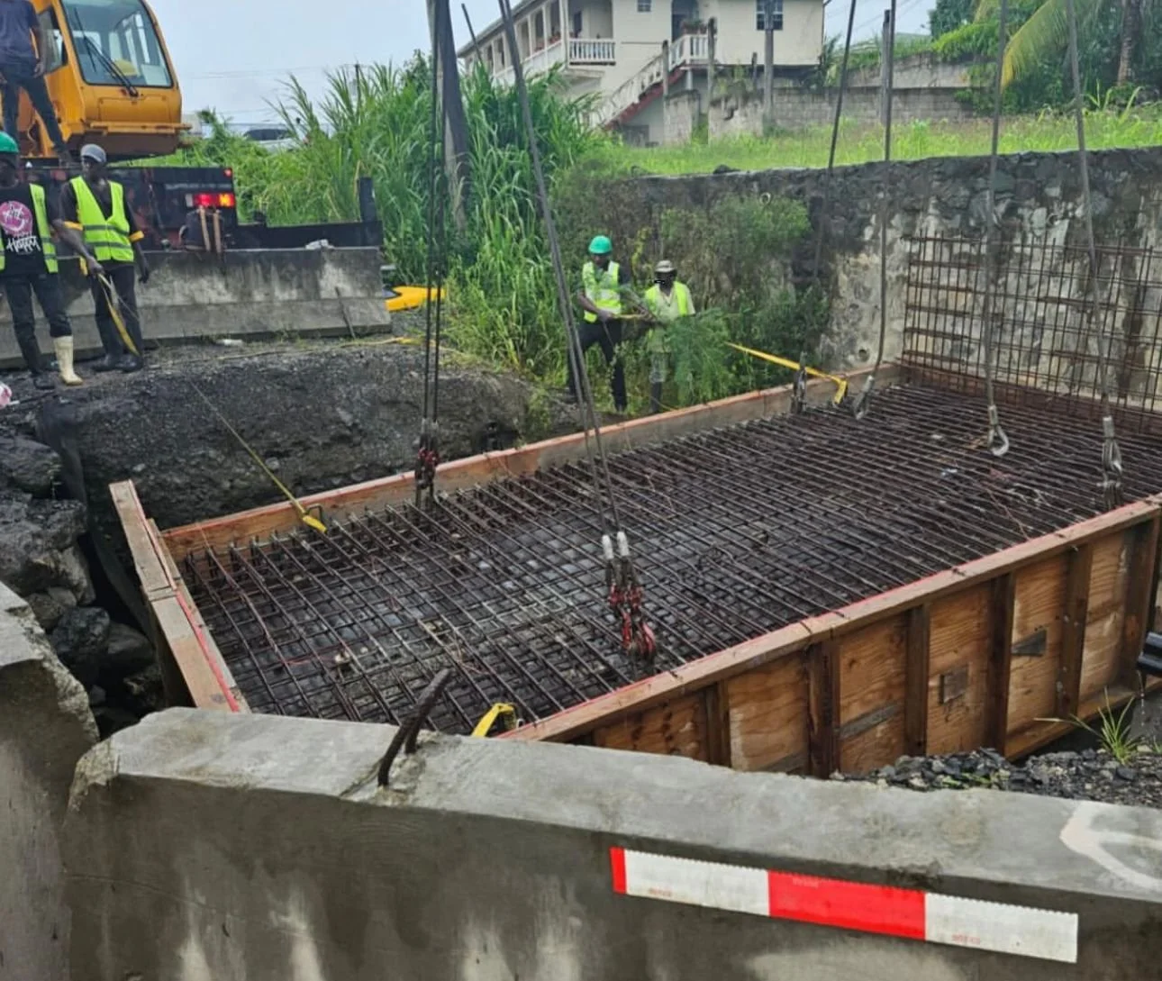 Construction workers in safety vests and helmets working on a building foundation with steel rebar reinforcement and concrete support structures.