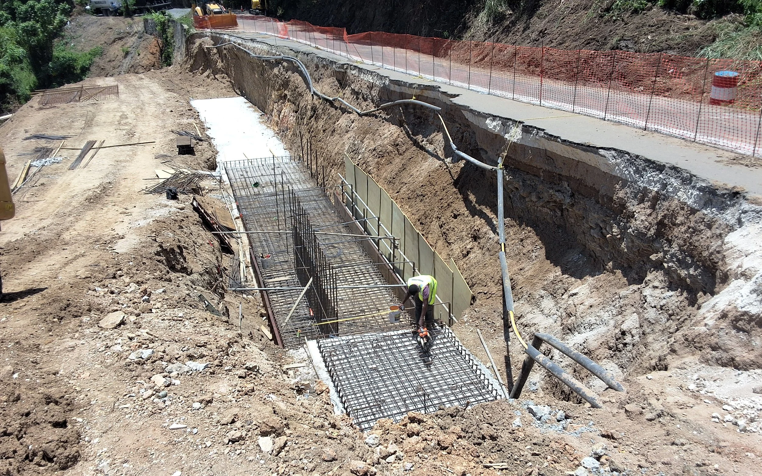 Construction worker installing steel reinforcement bars for a concrete structure in a deep trench on a hillside, with construction fencing and equipment visible.