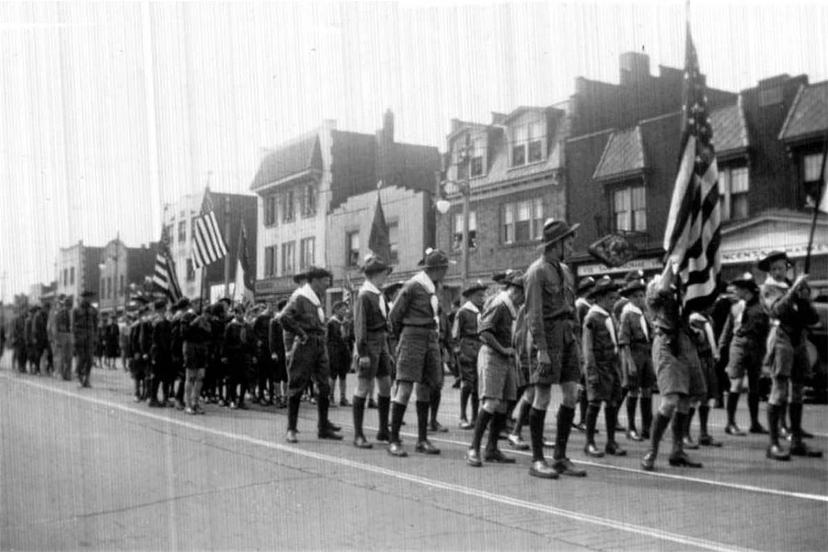 Boy scouts marching in a parade while holding American flags and a flag with stripes, in an urban neighborhood with Victorian-style buildings.