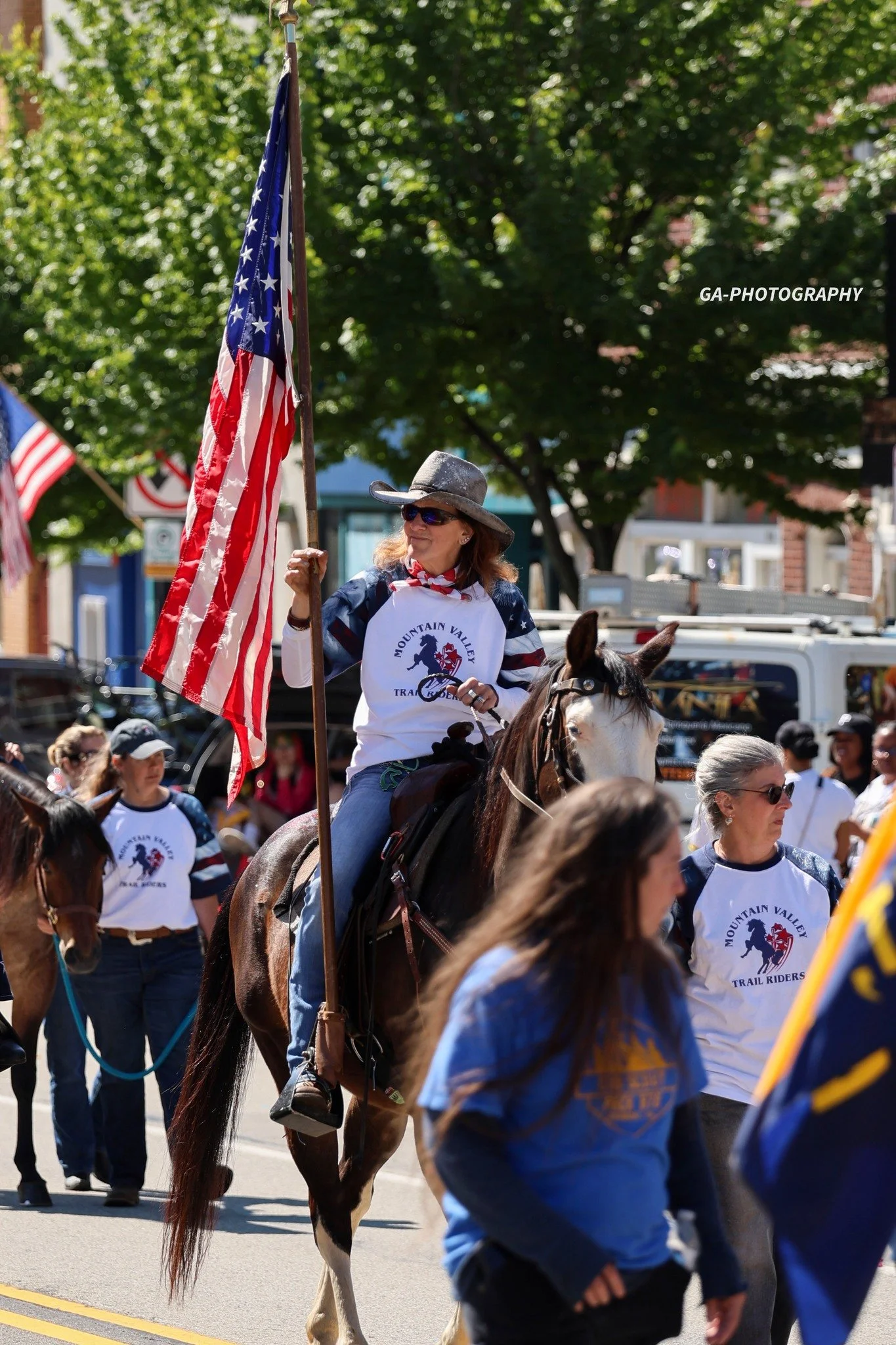 A woman riding a horse in a parade, holding an American flag, surrounded by other participants and spectators, some wearing shirts with 'Mountain Valley Trail Riders' logo.