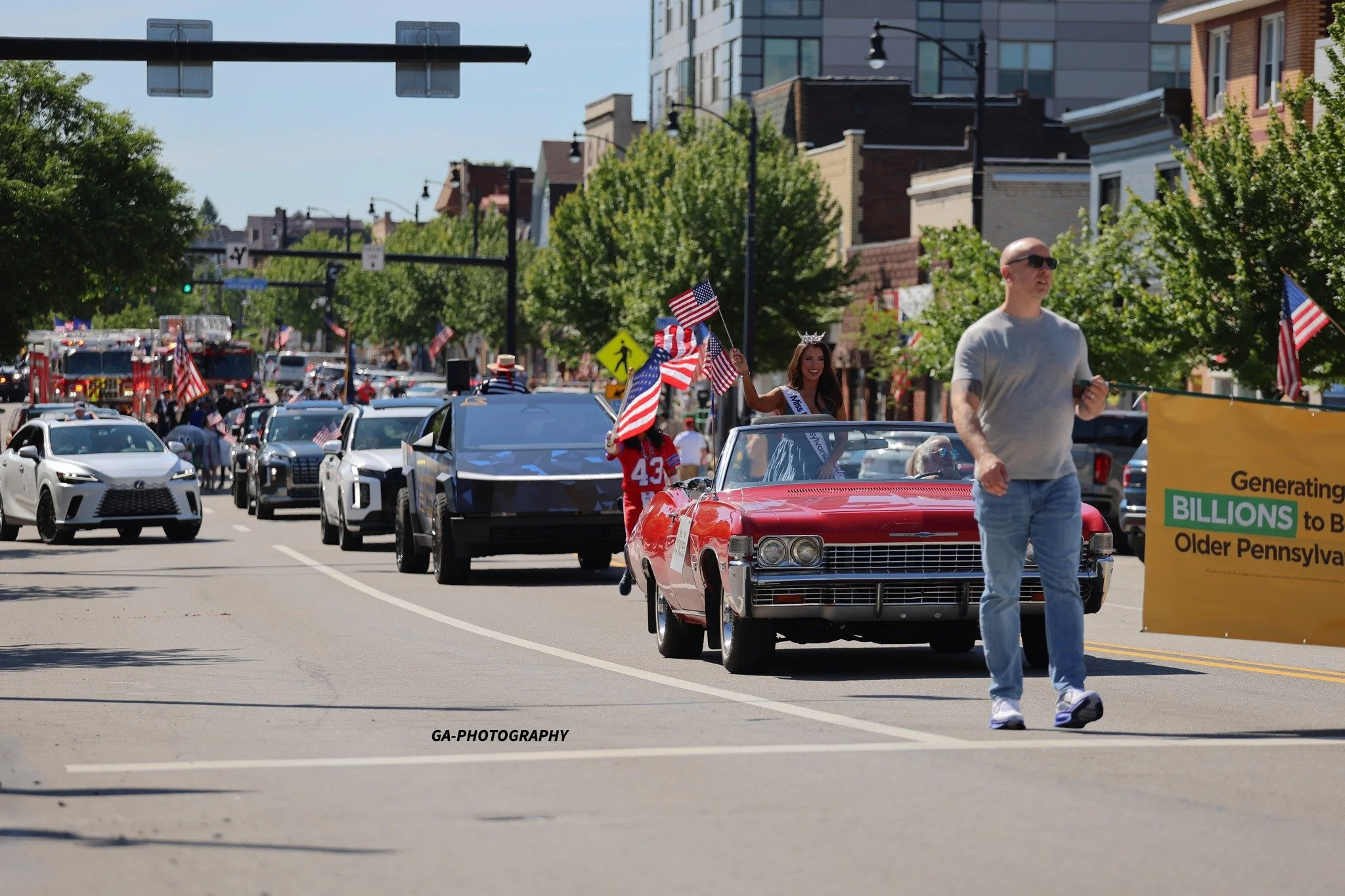 A parade on a city street with a woman wearing a crown and sash riding in a red convertible car, waving American flags, surrounded by cars and spectators, with a man walking beside the car holding a yellow banner. Trees line the street, and there are