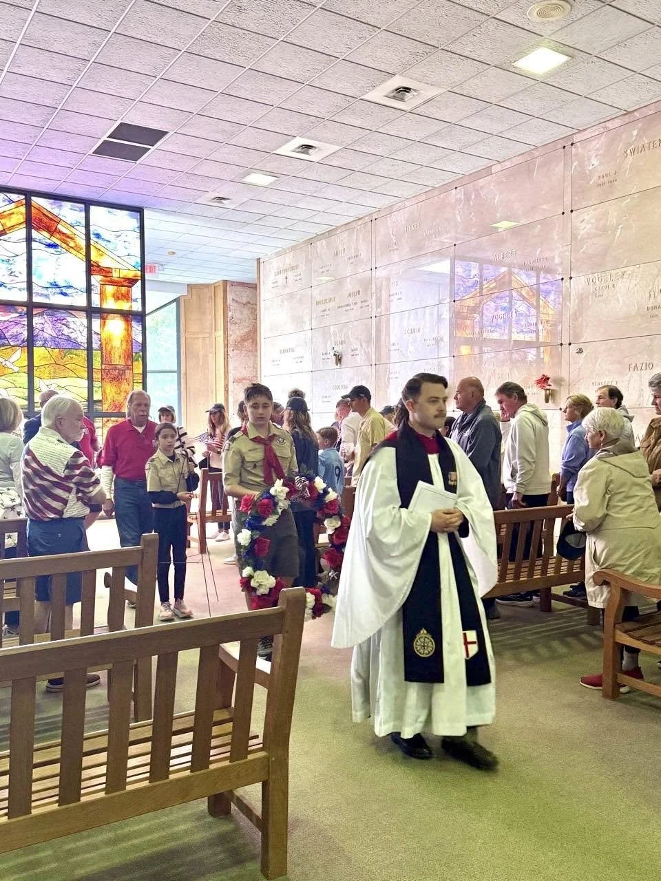 People gathered inside a church, with a priest in white robes standing in the foreground. Some children and adults are present, and there are floral wreaths and stained glass windows in the background.
