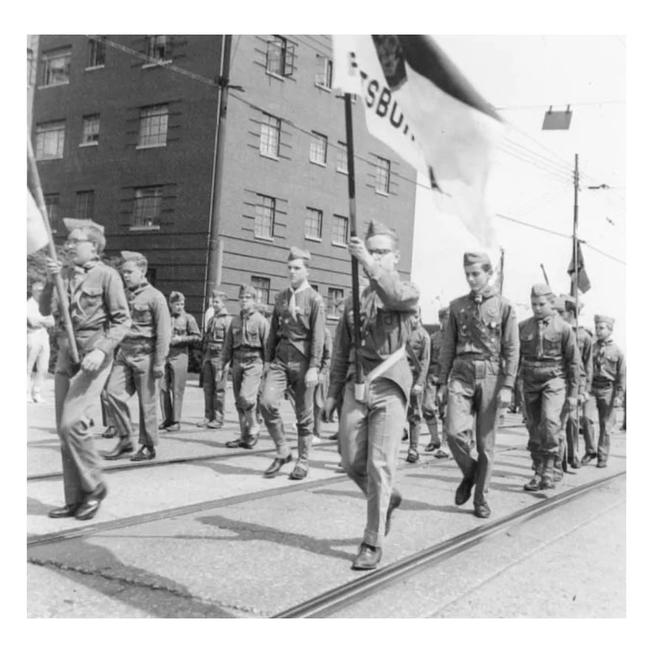Young boys marching in a parade, carrying flags, in front of a building, in what appears to be a historical black-and-white photo.
