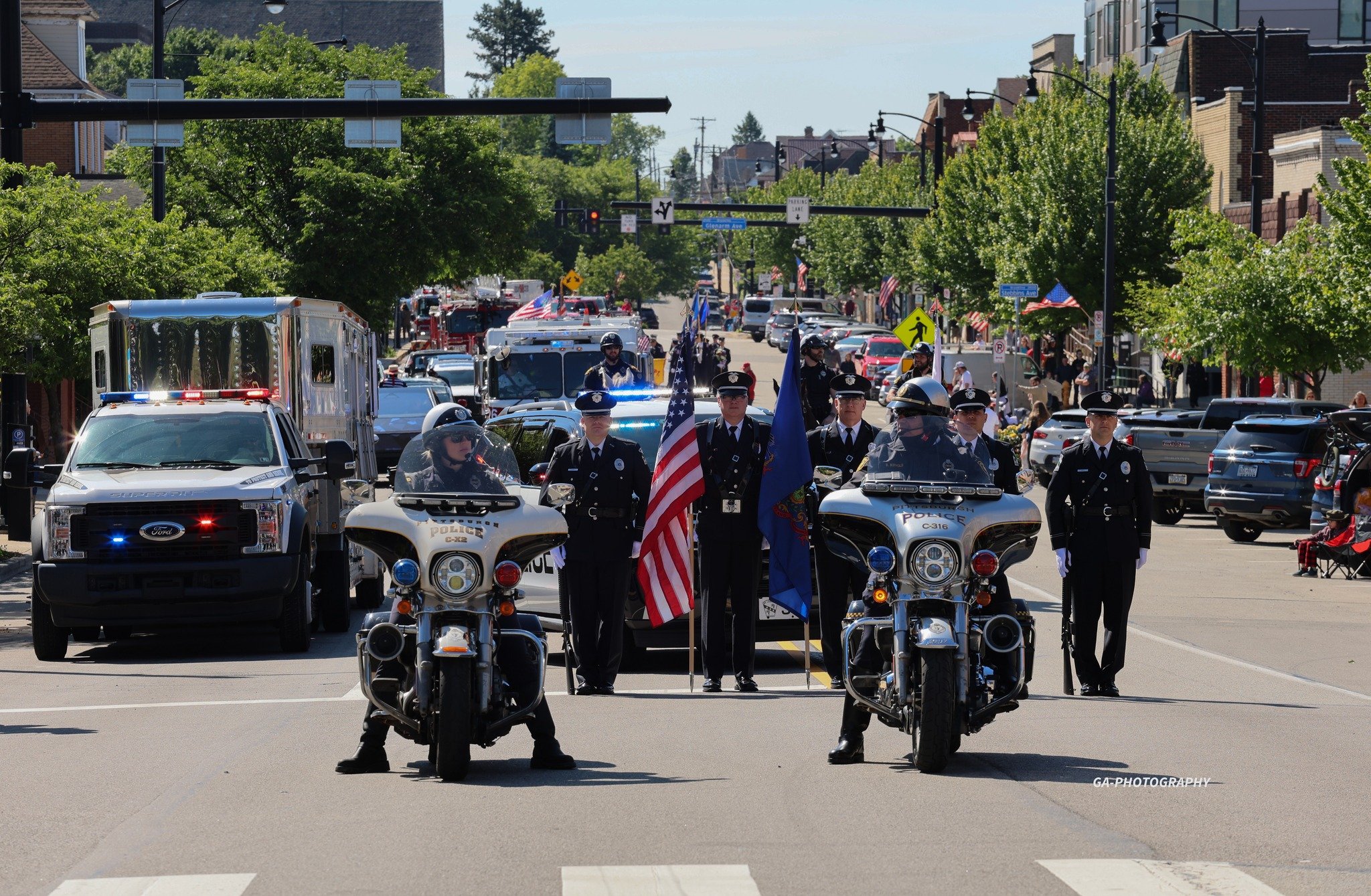 Law enforcement officers participating in a parade, with two officers on motorcycles at the front, holding American flags and other flags, in a town street lined with parked cars, trees, and spectators.