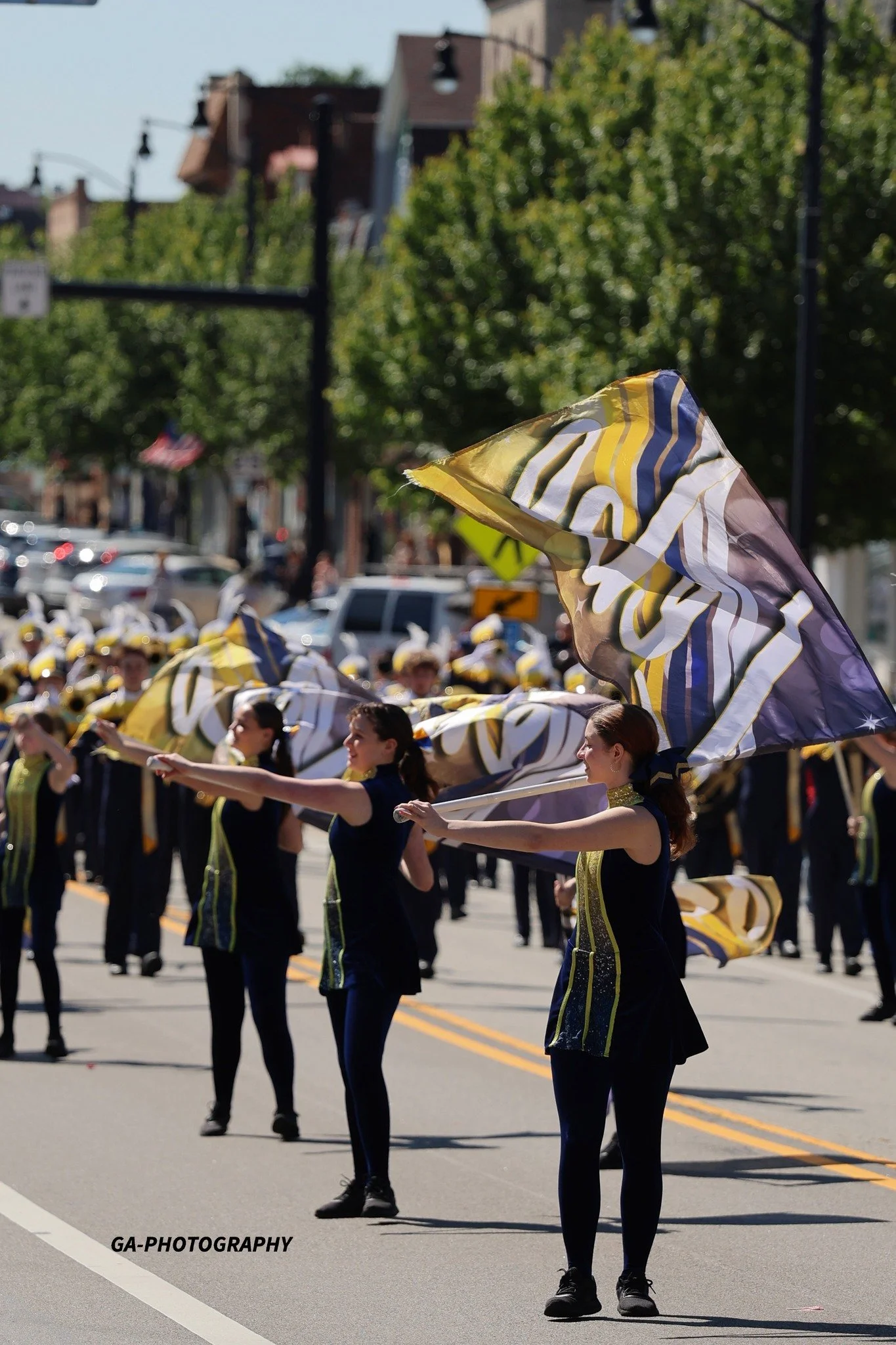 Group of women marching in a parade, waving large yellow, white, and blue flags, dressed in dark blue outfits with yellow accents, on a sunny street lined with cars and trees.