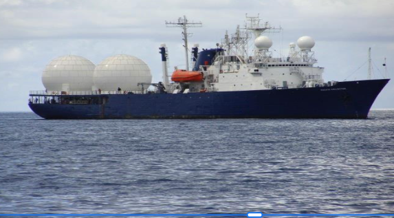 Large military ship with radar domes and communication equipment sailing in the ocean.