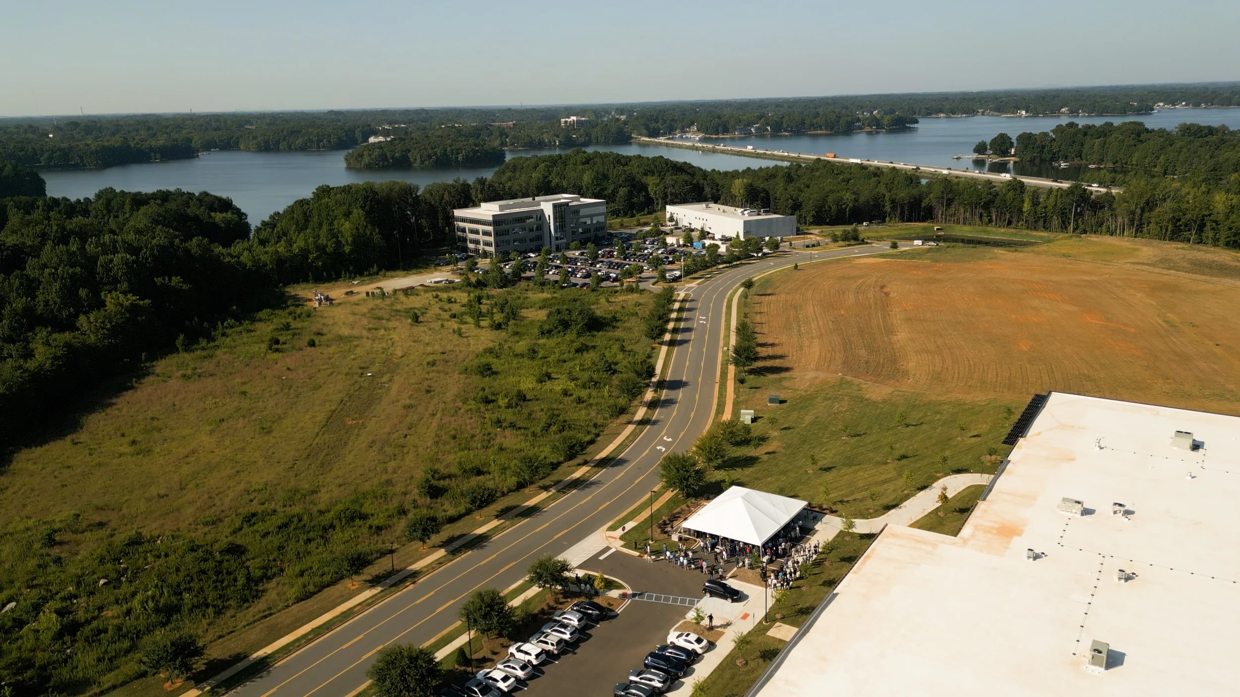 An aerial view of a park with a road, parking lot, white tent, surrounding green trees, open fields, and a large lake in the background.