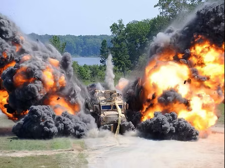 Explosion with fire and smoke, debris, and flames engulfing a vehicle on a road near a body of water and trees.
