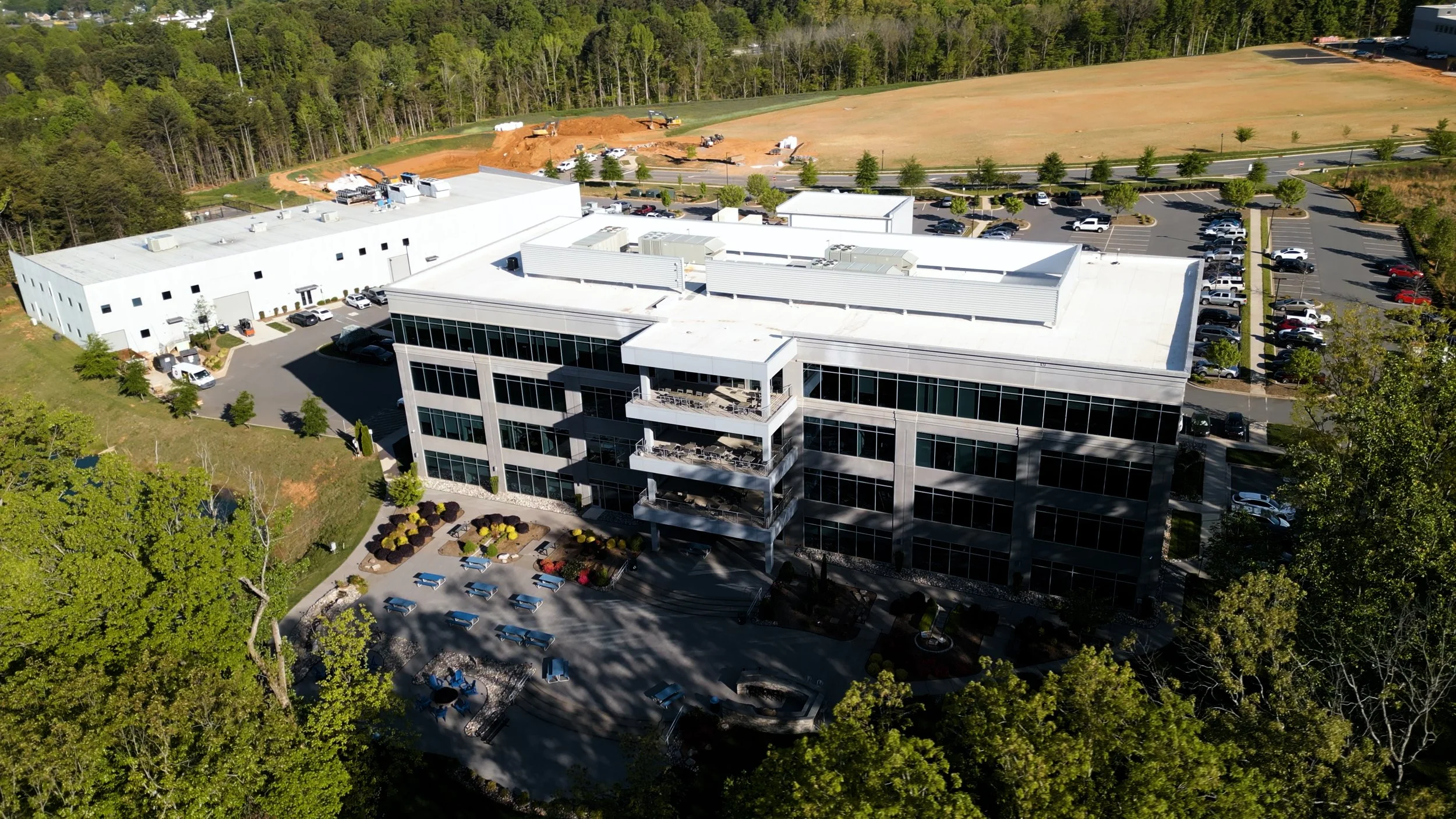 Aerial view of a modern office building with glass windows, surrounded by parking lots with numerous cars, landscaped gardens, and trees, with a construction site and wooded area in the background.