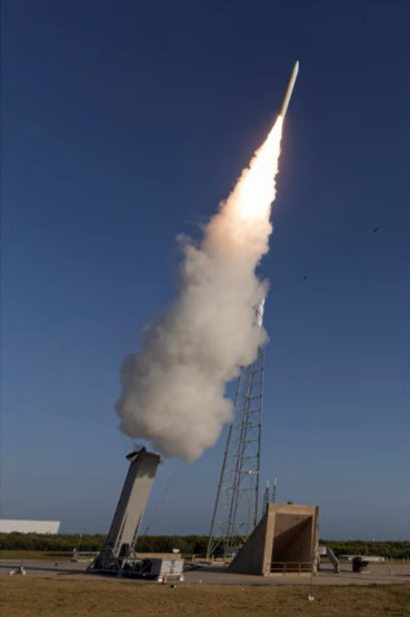 Rocket launching from a platform with smoke and fire against a clear blue sky.