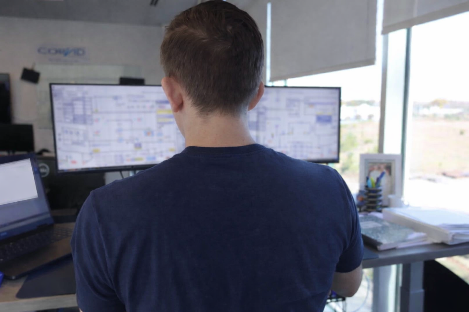 A person with short brown hair working at a desk with multiple computer screens displaying complex schedules or diagrams, in an office space with large windows and blinds.