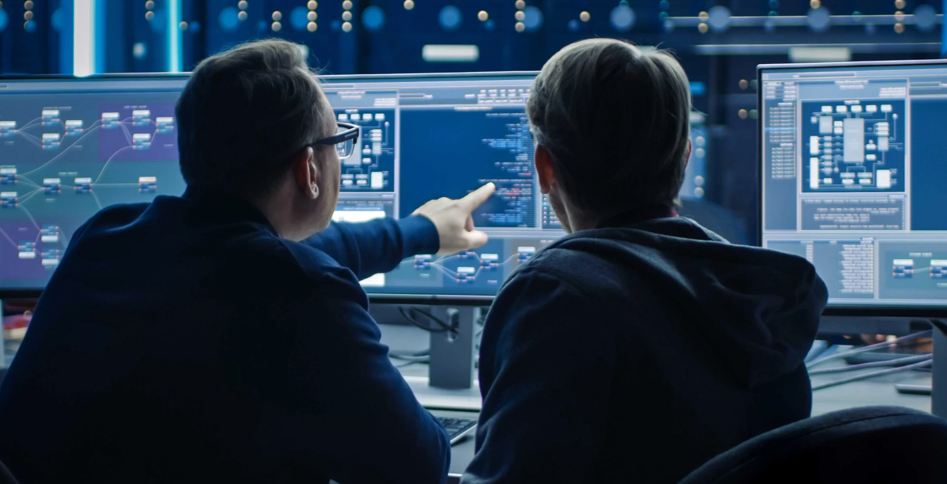 Two people working on multiple computer screens displaying complex data and diagrams in a dimly lit room.