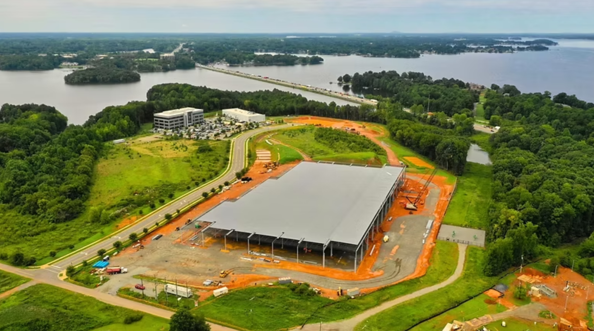 Aerial view of a construction site with a large building under construction, surrounded by greenery, near a body of water in the background.