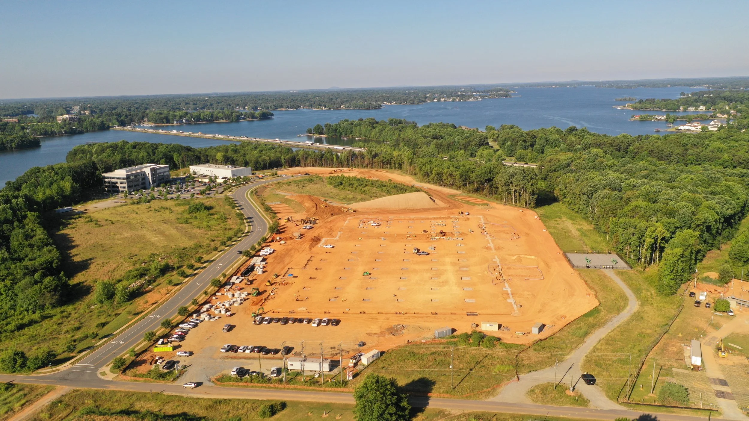 A construction site with cleared and leveled land surrounded by trees, near a river with a bridge, and a building complex with parking lot.